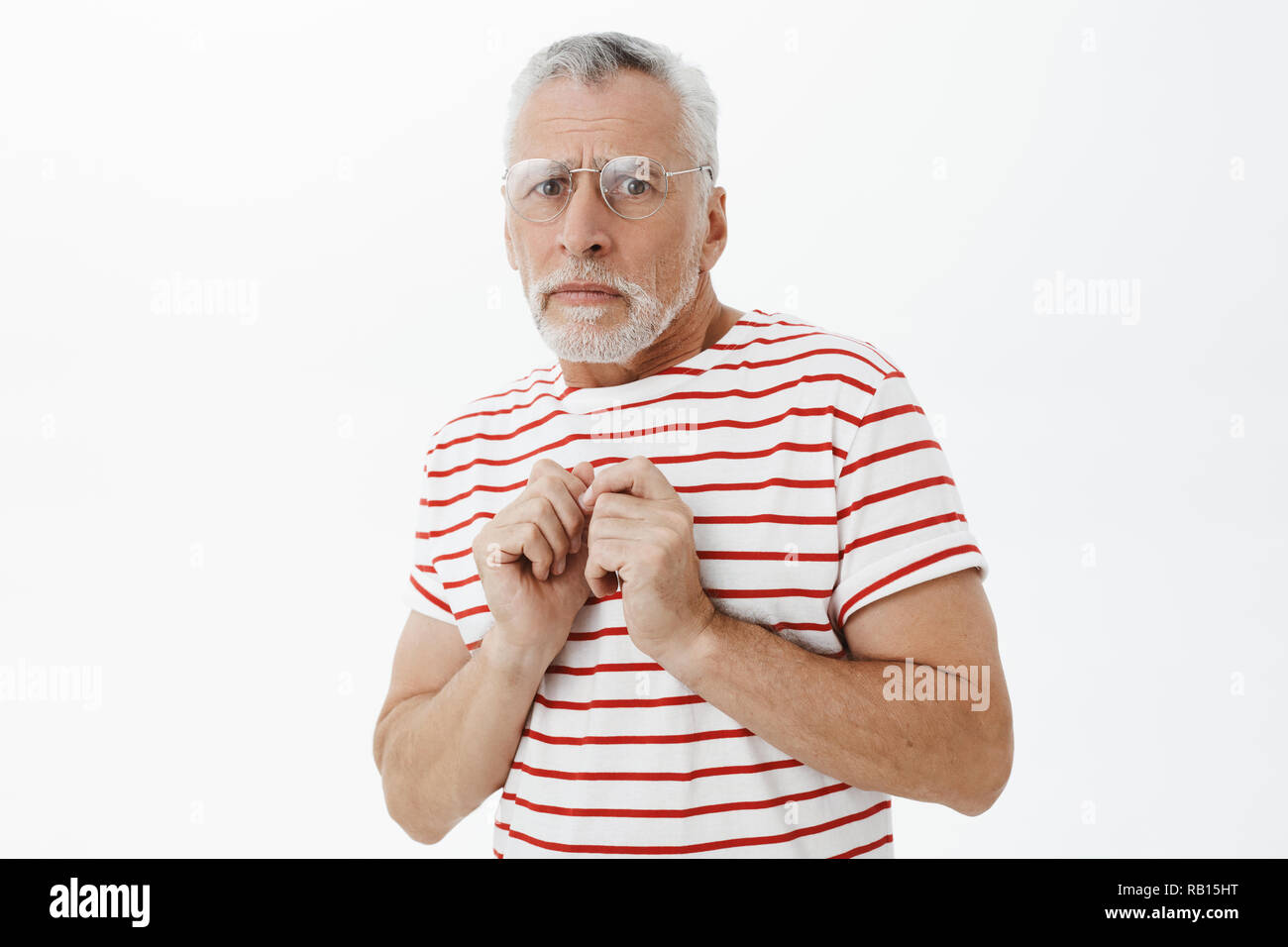 Timid and insecure senior man in striped t-shirt with grey hair and beard pressing hands to body from fear and anxiety making nervous scared expression being lonely and not able to stand Timid and insecure senior man in striped t-shirt with grey hair and beard pressing hands to body from fear and anxiety making nervous scared expression being lonely and not able to stand