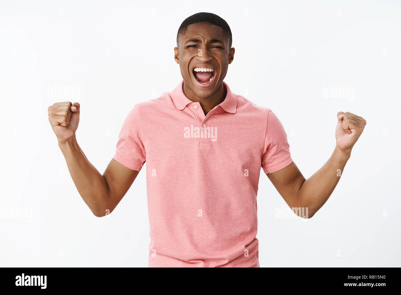 Guy releasing regret and negative emotions after seeing failure clenching raised fists screaming and frowning looking upset and unhappy after favorite team losing, standing sad over gray background Stock Photo