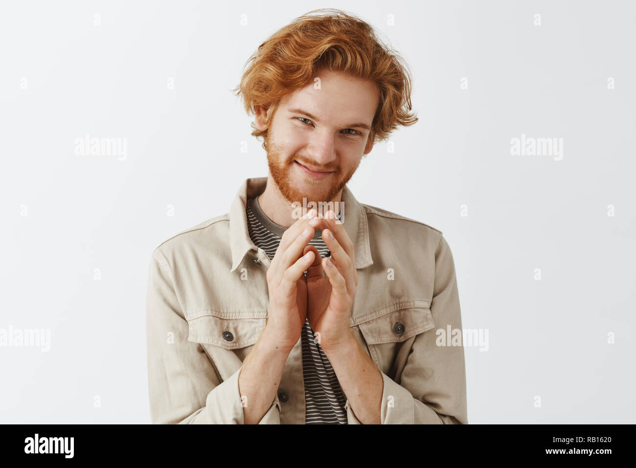 Redhead bearded man has tricky and evil plan on mind steepling fingers over chest and smirking mysteriously squinting at camera standing in beige jacket over gray background. Evil genius in action Stock Photo