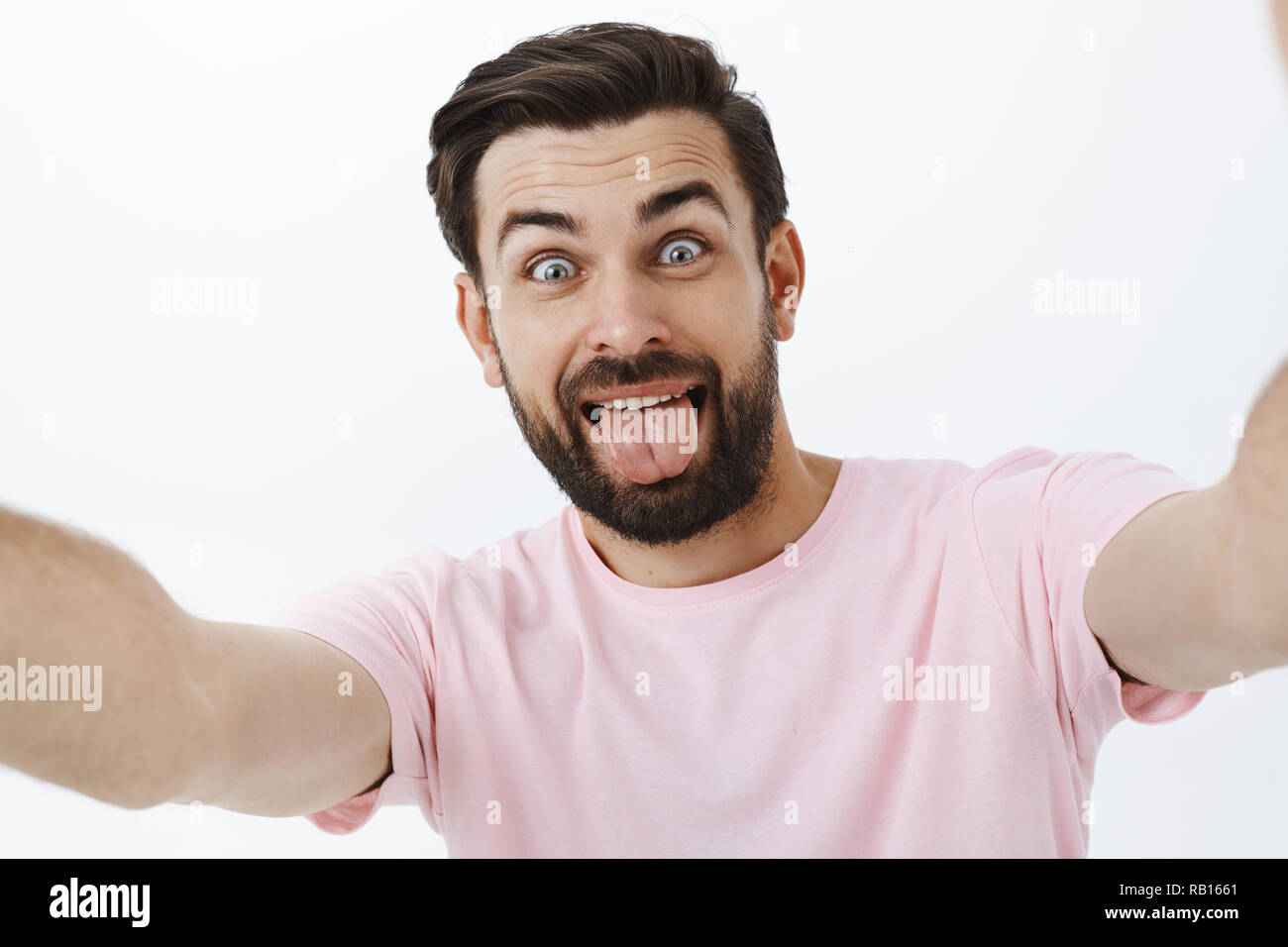 Cute and carefree adult bearded man with blue eyes and beard having fun, fooling around holding camera with extended hands sticking out tongue and popping eyes, aping playfully from joy Stock Photo