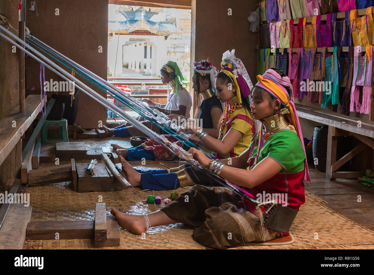 Four Kayan (Padaung) women wearing brass neck coils and colourful traditional dress, weaving cloth using traditional looms with a pagoda in the backgr Stock Photo