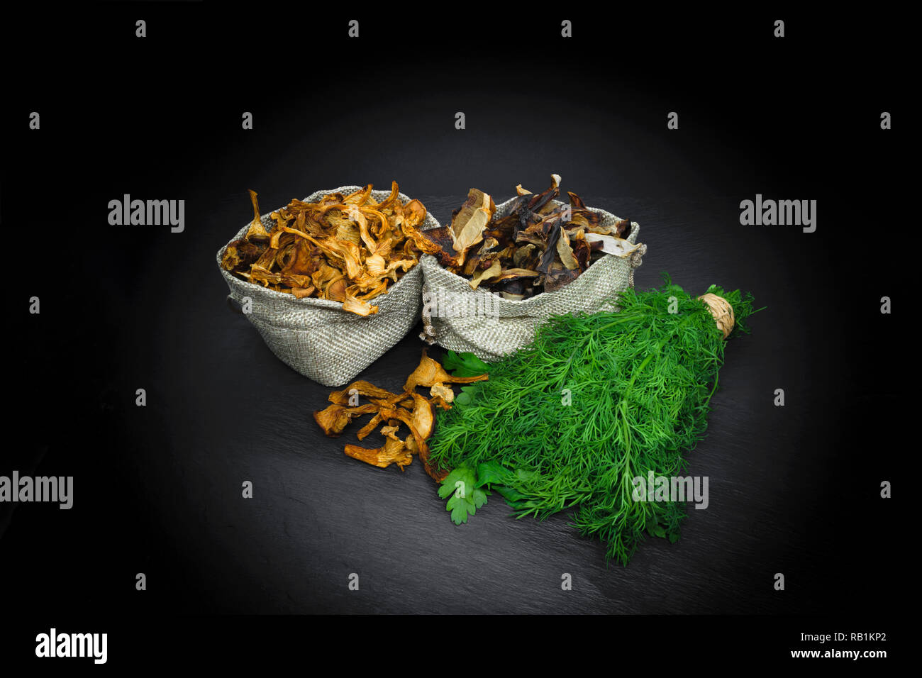Composition of dry boletus and chanterelles mushrooms placed in canvas bags with dill and parsley broom on black stone background surface with copy sp Stock Photo