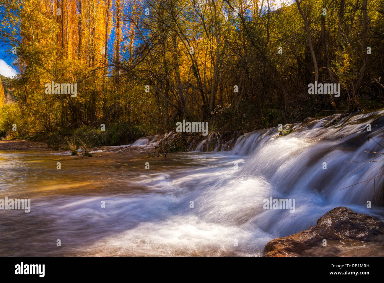 Low angle photo of a small waterfall on a crisp autumn day with golden yellow and orange leaves on the trees of the surrounding forest and blue sky Stock Photo