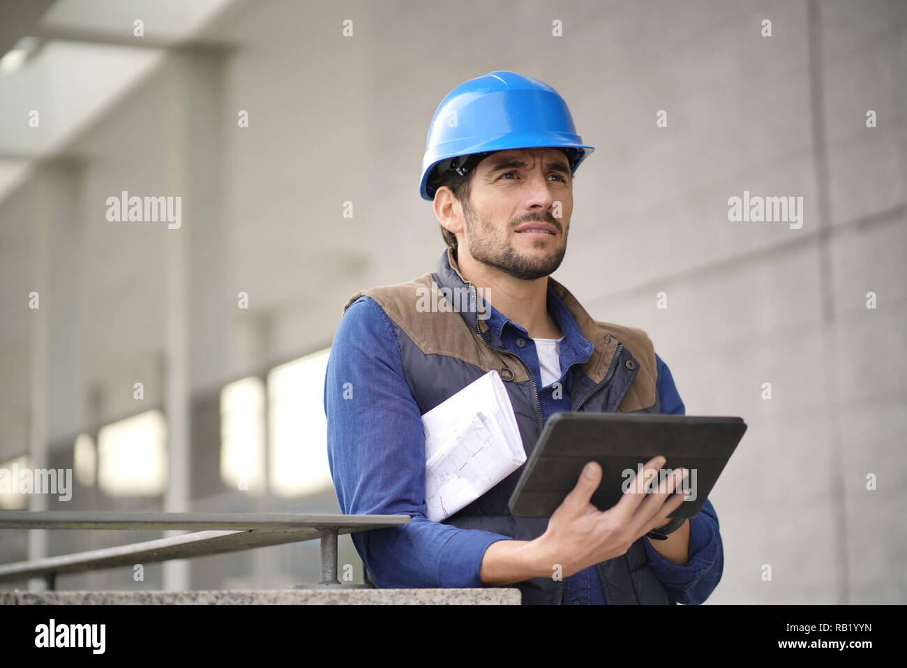 Handsome building expert in hardhat outdoors with tablet and blueprint ...