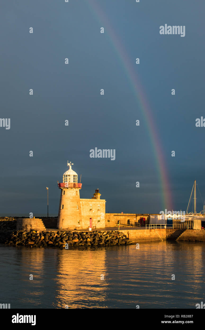 The lighthouse at Howth Harbour in County Dublin bathed in sunlight under a rainbow in front of a stormy sky, with its reflection in the water. Stock Photo