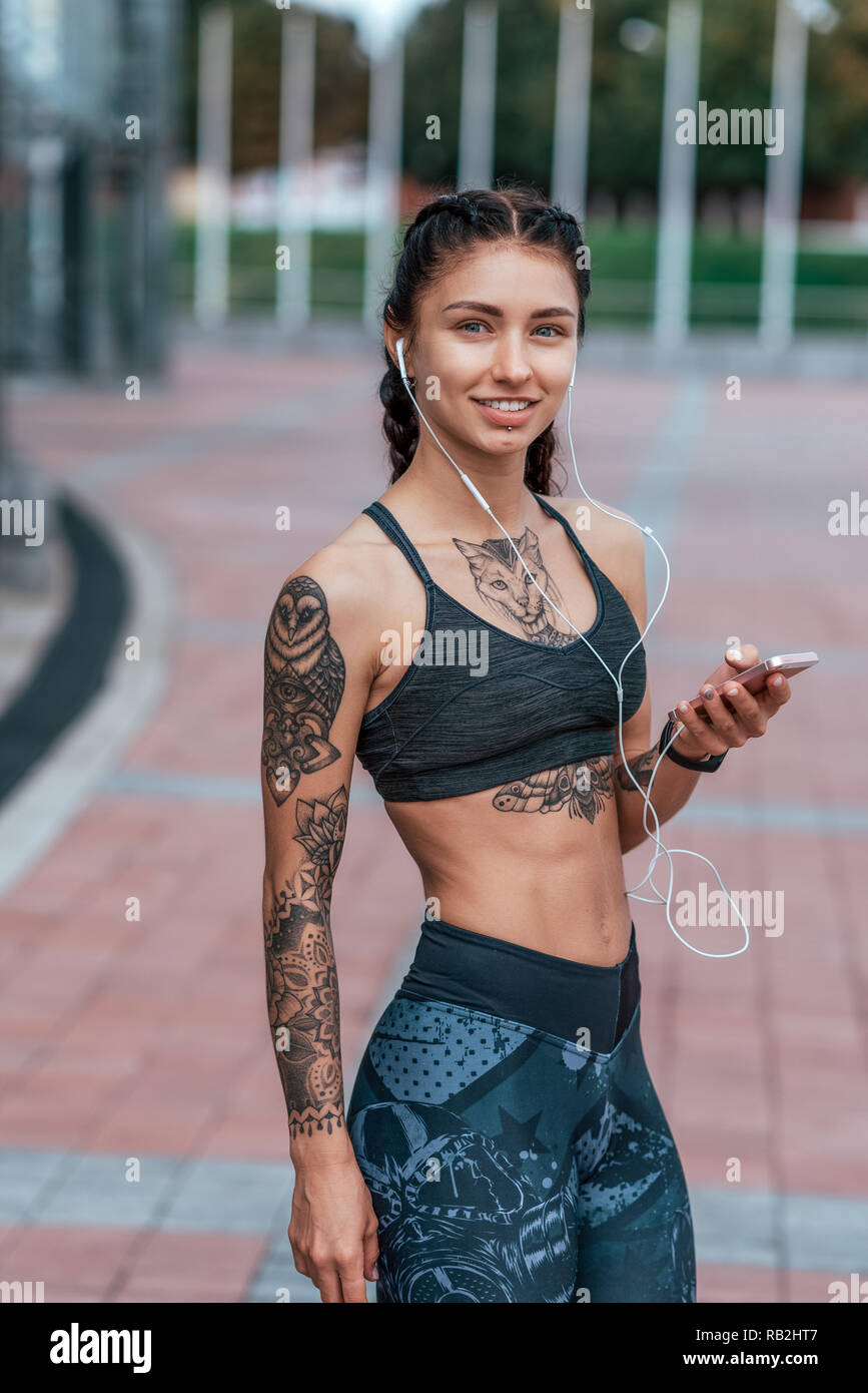 Girl in sportswear. In his hand, smartphone listens music headphones.  Posing summer morning in city. Beautiful Tattoos tanned female skin. Happy  smiling, relaxing after hard workout Stock Photo - Alamy, image size:867x1390
