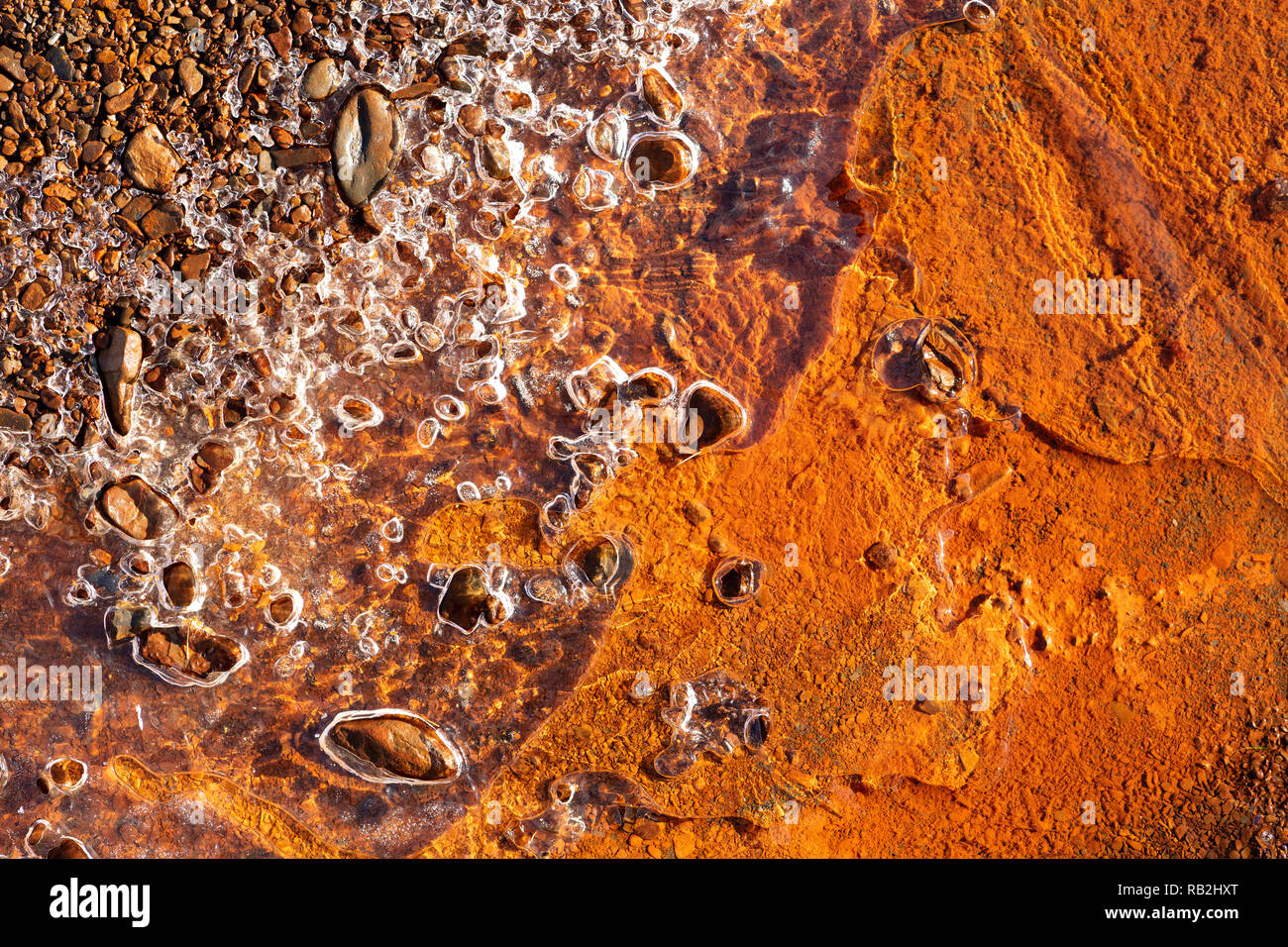 rusty orange background under a sheet of frozen water, some stones protude from the transparent ice sheet Stock Photo