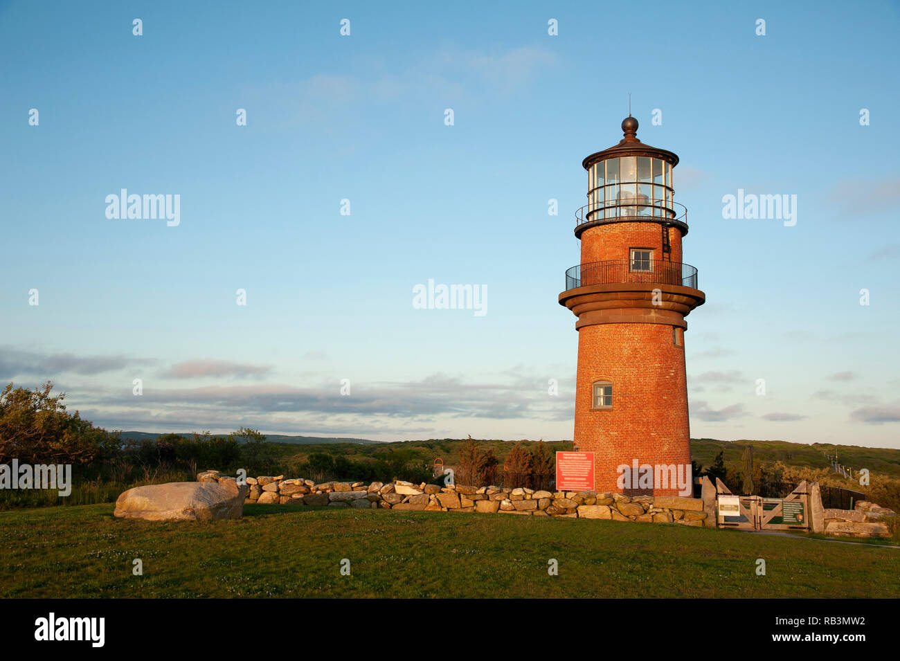 Aquinnah lighthouse, also referred to as Gay Head light, sits on a hilltop, surrounded by a stone wall and wooden gate as the sun sets on Martha’s Vin Stock Photo