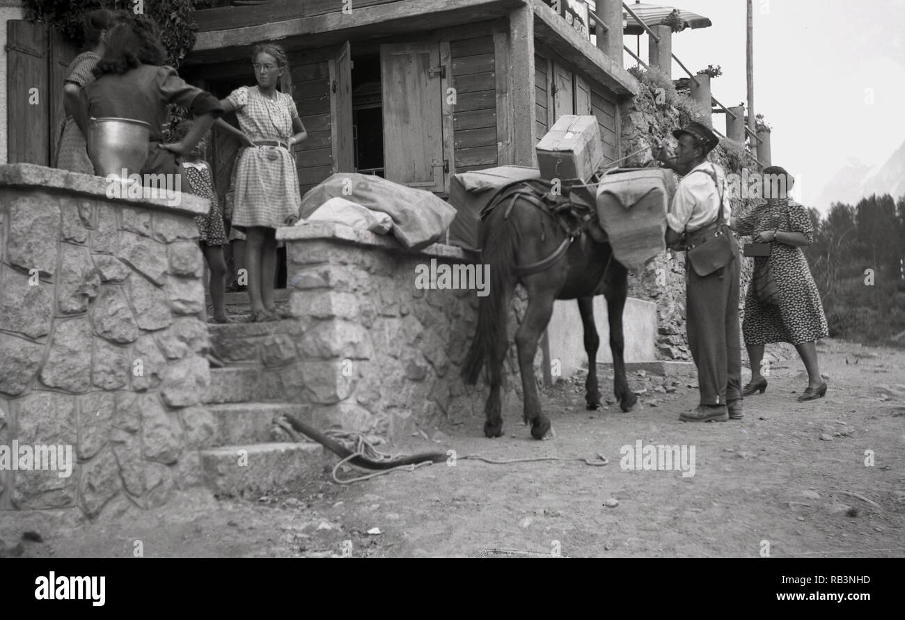1950s, historical, a postman on a dusty track with a donkey carrying mail, delivering parcels to a wooden chalet in the mountains, Switzerland. Stock Photo