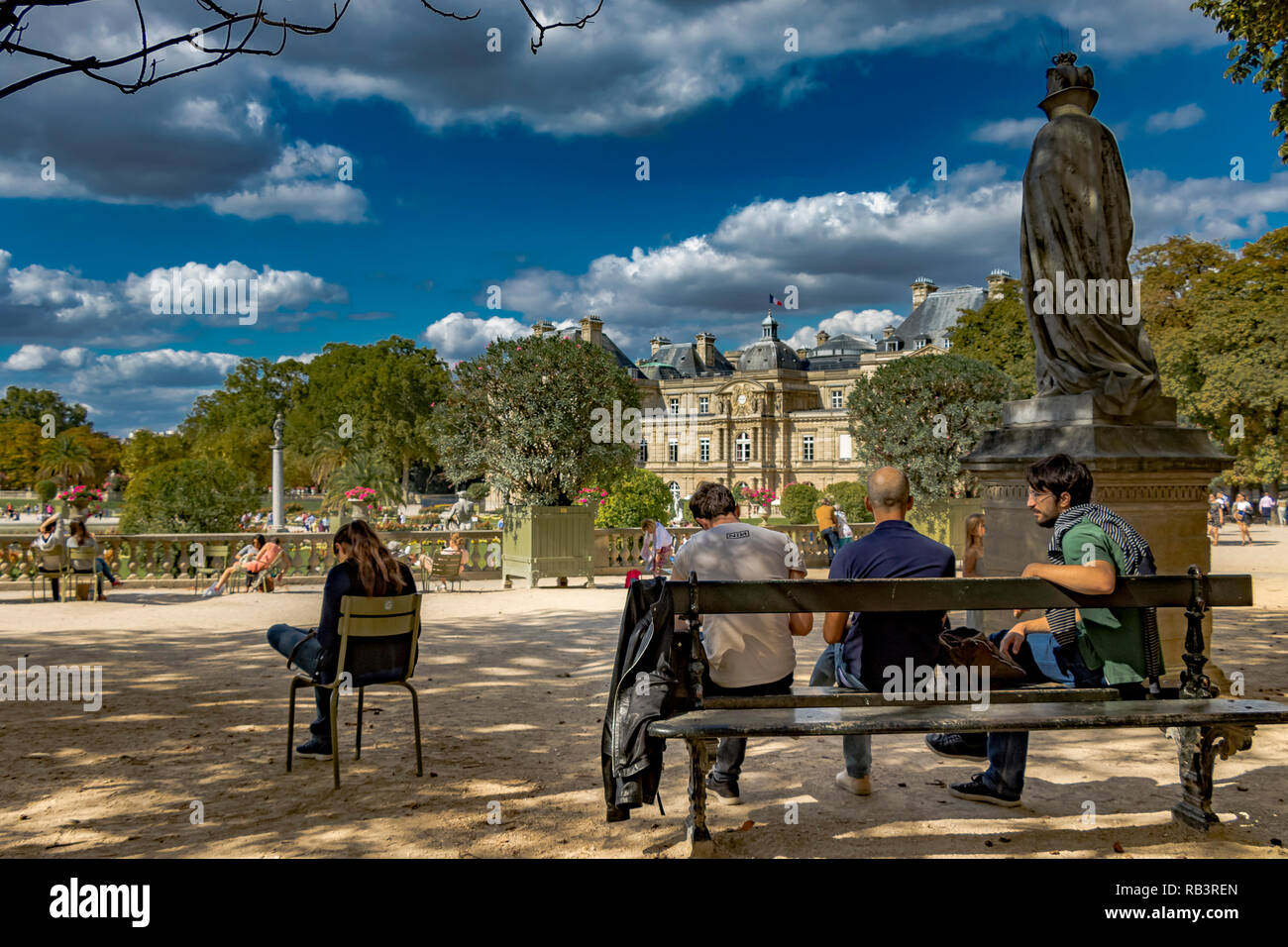 A girl sits on a green chair checking her phone whist nearby three men sit on a bench relaxing on a summer's day in the Jardin du Luxembourg,Paris, Stock Photo