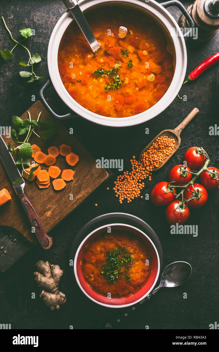 Lentil soup in pot and bowl with spoon and ladle. Cooking ingredients on dark rustic kitchen table background, top view. Healthy vegan food concept. V Stock Photo