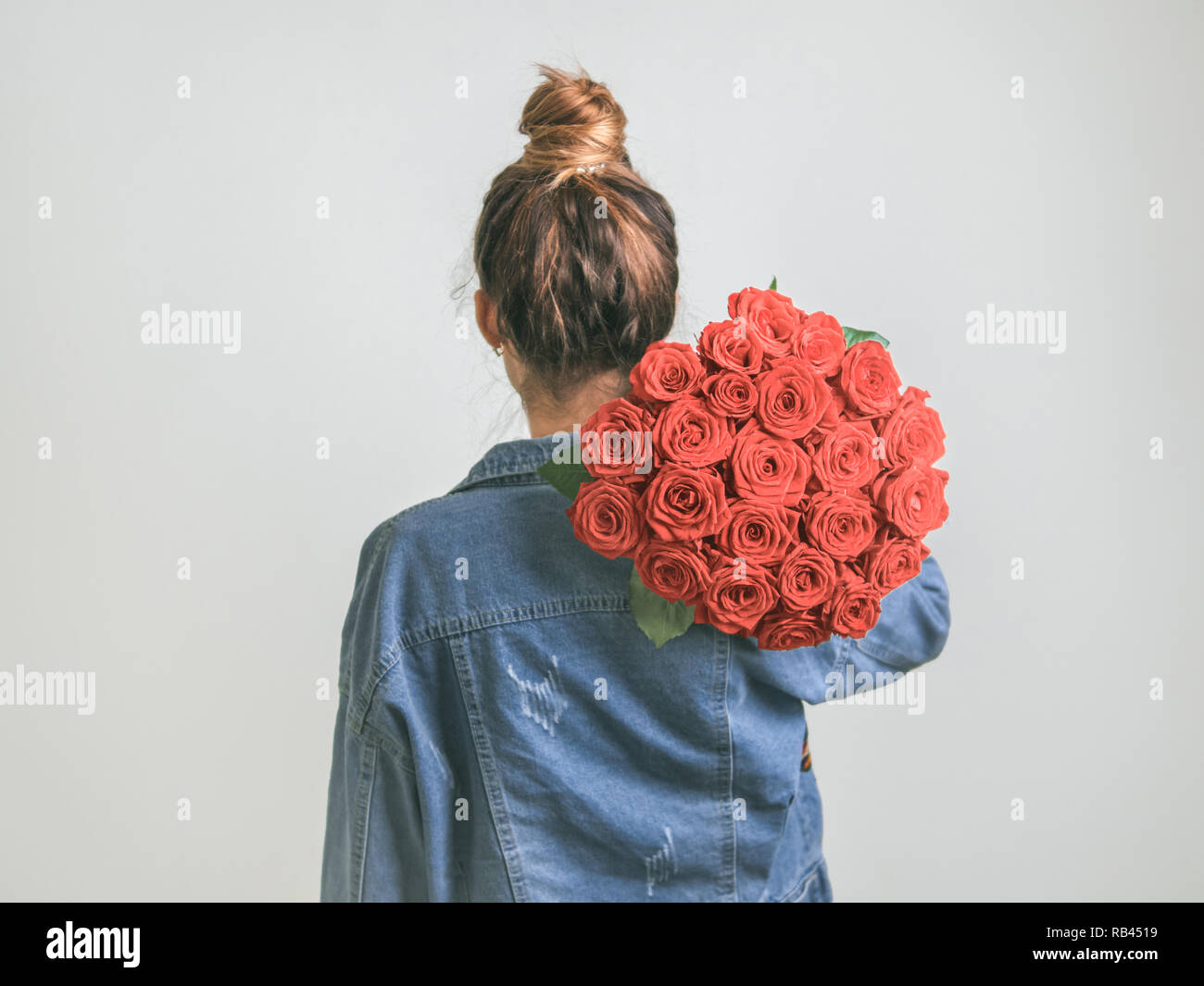 Back view of young woman in denim jacket holding bunch of Living Coral roses on shoulder. Girl with bun updo in jeans holding flowers in Color of Year Stock Photo