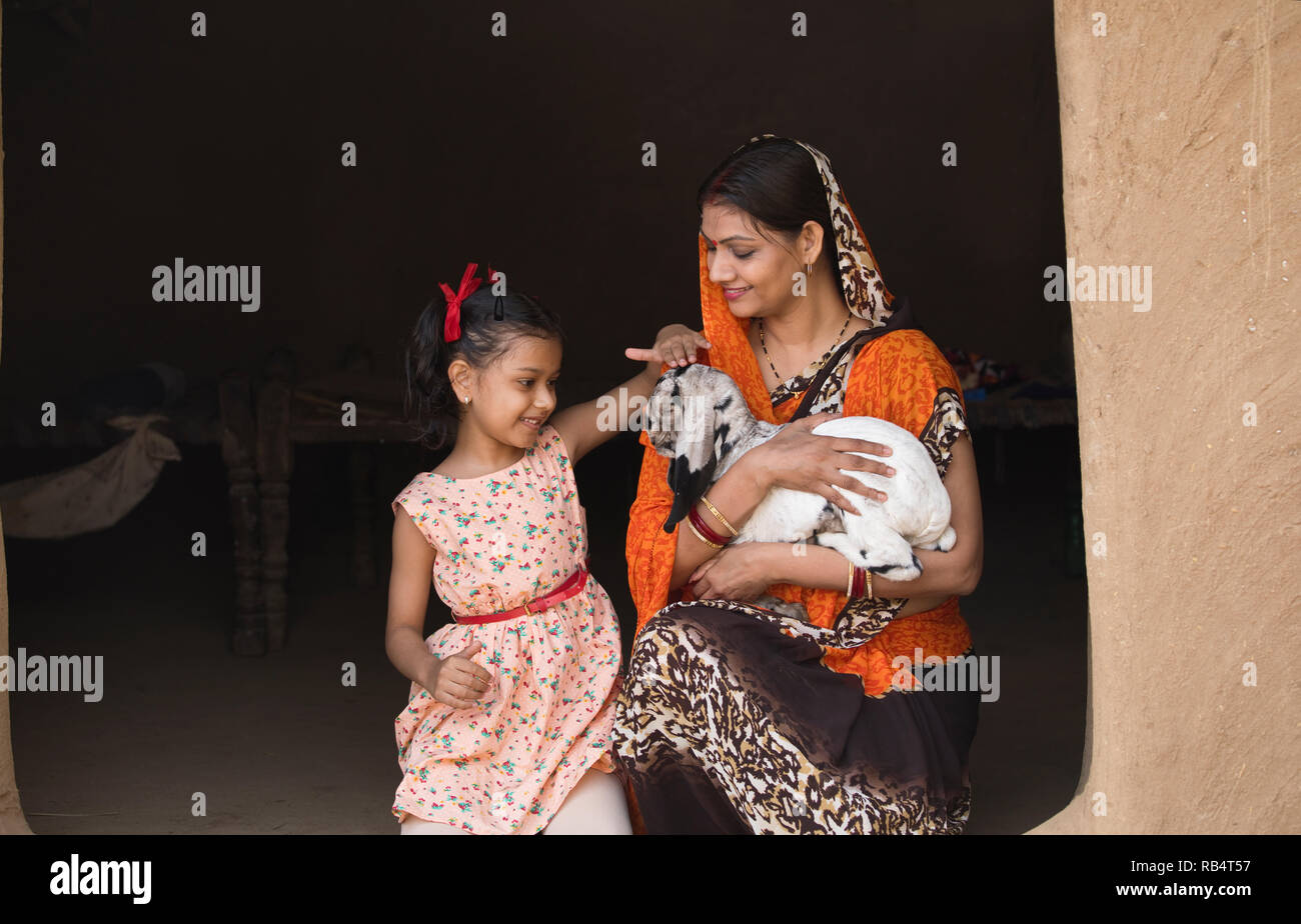 Happy Indian mother with her daughter stroking baby goat at village house Stock Photo