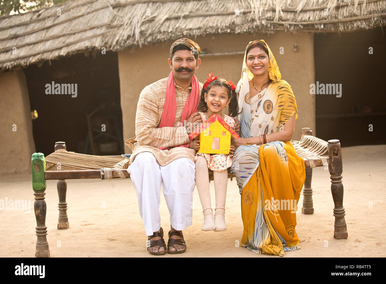 Rural Indian family holding dream house model while sitting on traditional bed in village Stock Photo