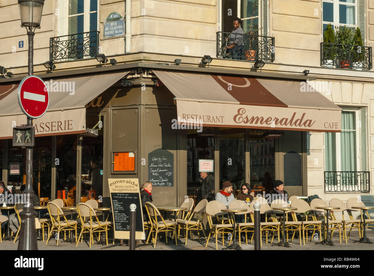People sat drinking coffee outside Parisian restaurant 'Esmerelda' beside Notre Dame Cathedral, in spring sunshine with a women at the balcony above. Stock Photo