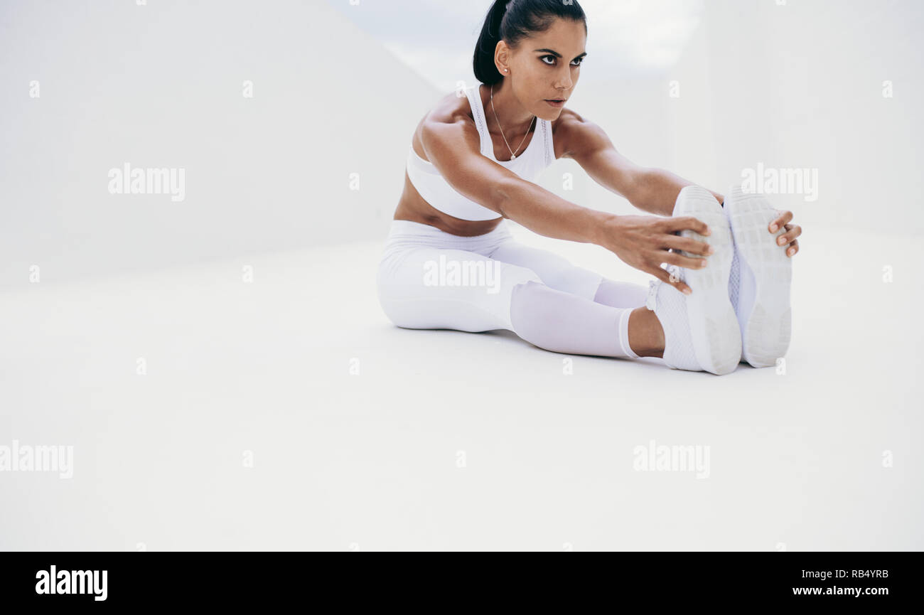 fitness woman doing stretching exercises. Woman in white fitness wear sitting on floor and bending forward to hold her legs. Stock Photo