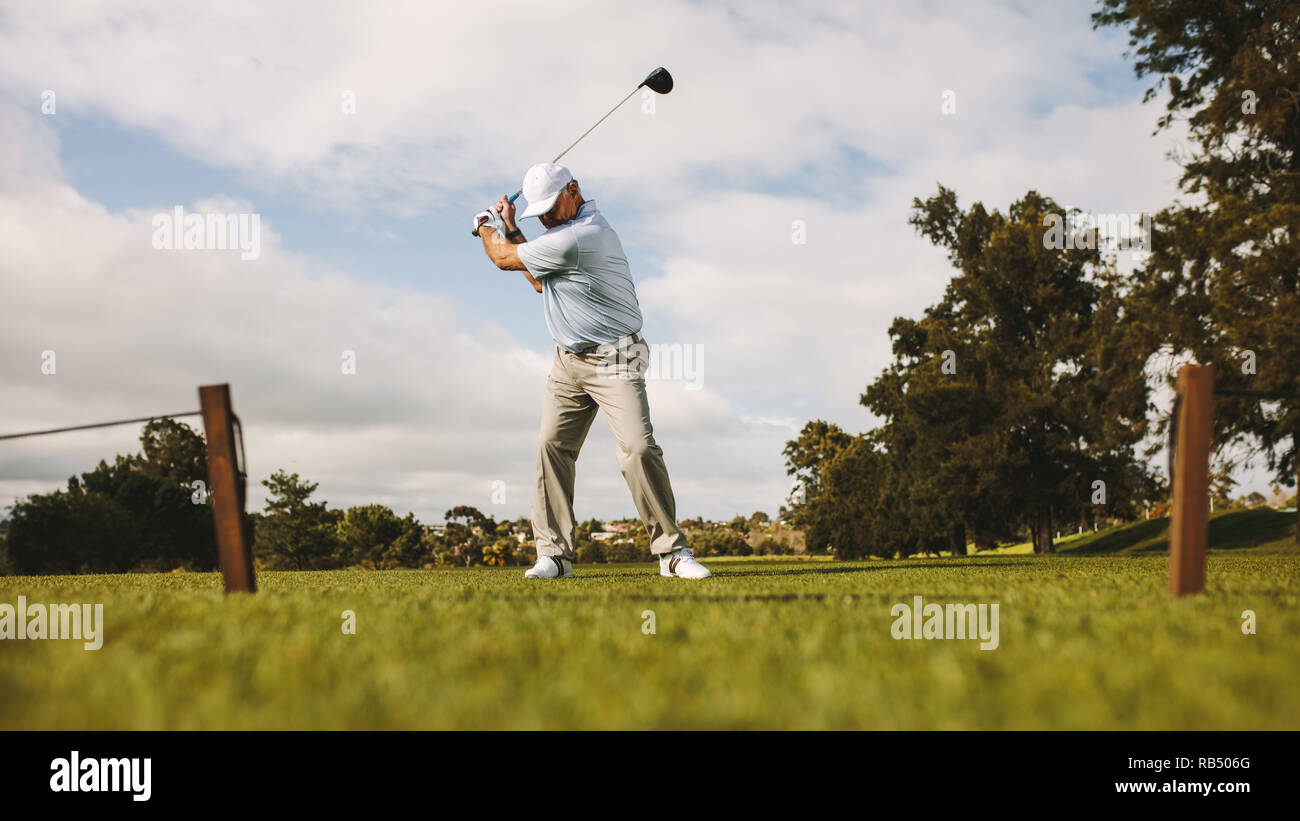 Low angle shot of senior male golfer taking shot while standing on field. Full length of golf player swinging golf club. Stock Photo