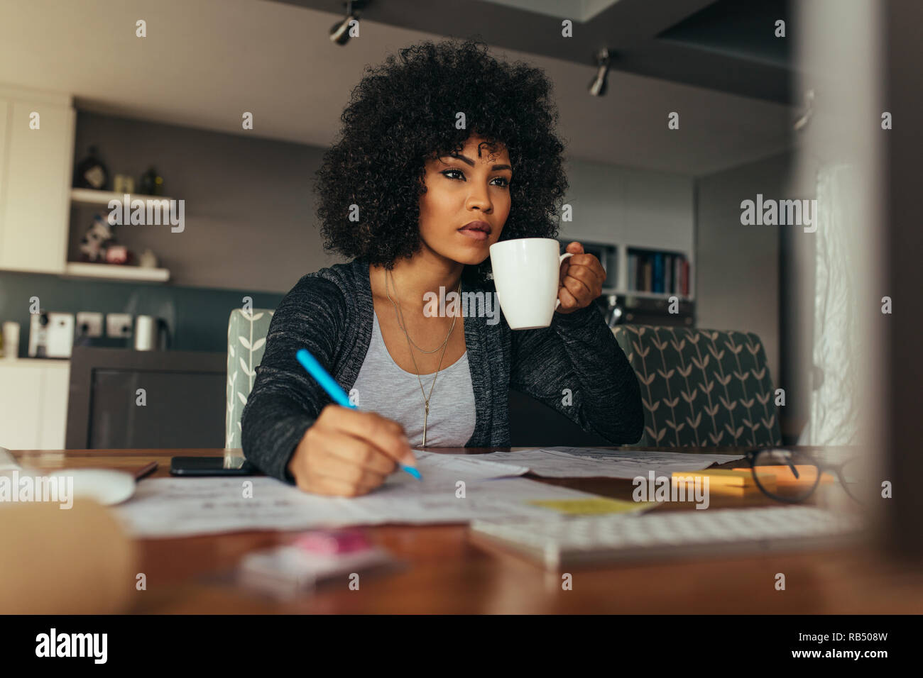 African woman working at her desk making notes looking at computer monitor and drinking coffee. Female architect working at her home office desk. Stock Photo