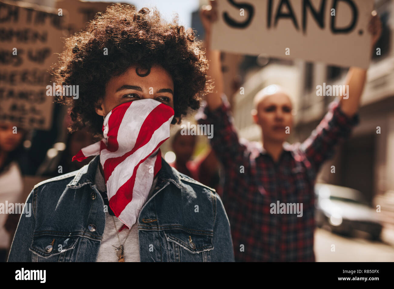 Portrait of silent protesting young woman with group in background. African female covering her mouth with a scarf and protesting on road with group o Stock Photo
