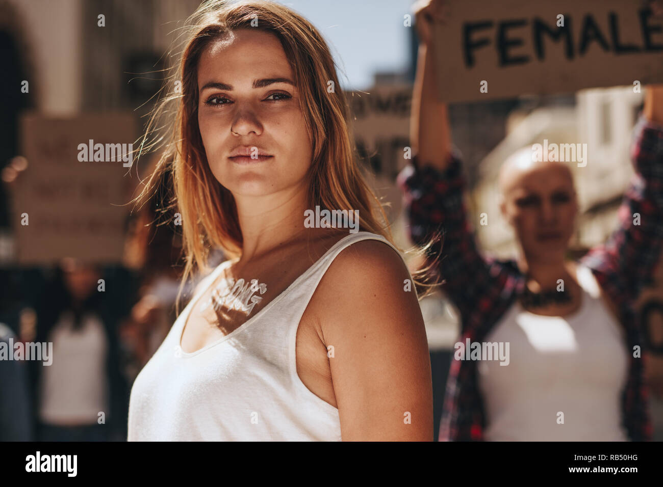 Woman activist with word strong written on her body standing outdoors. Strong young female activist protesting outdoors with her group in background. Stock Photo