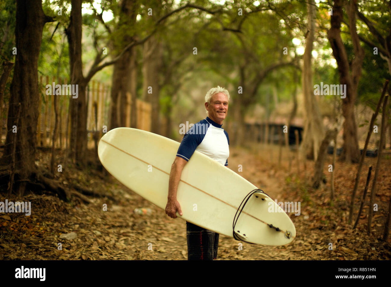 Mature man smiles as he stands on a rural path carrying a surfboard and poses for a portrait. Stock Photo