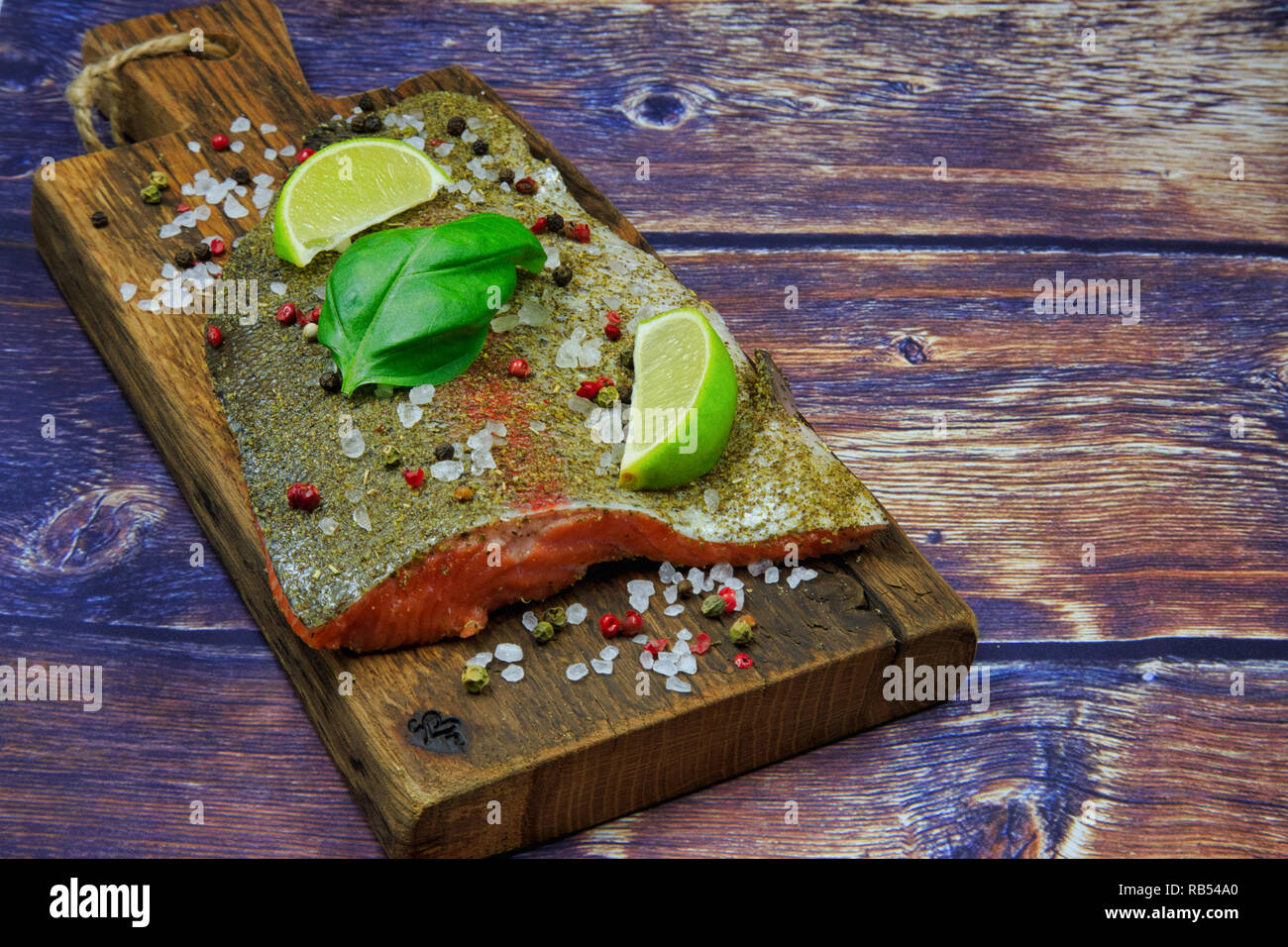 Raw rainbow trout steak sprinkled with spices, pepper, salt with pieces of lime and basil leaves on an old wooden board. Flat view from above in vinta Stock Photo