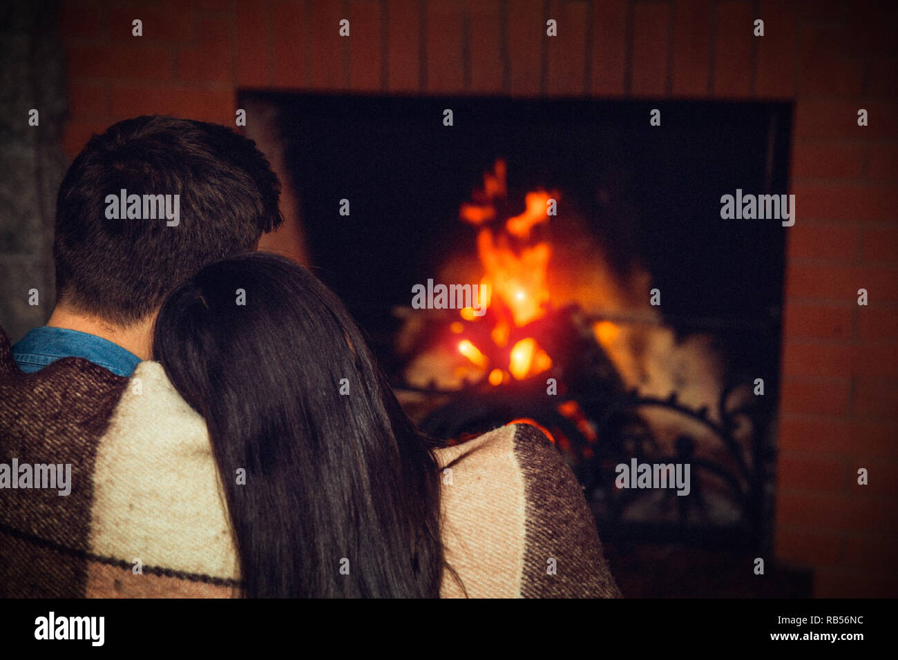 Brunette young man and woman sit together and look at fireplace. She hold her head on his shoulder. They covered with blanket. Stock Photo