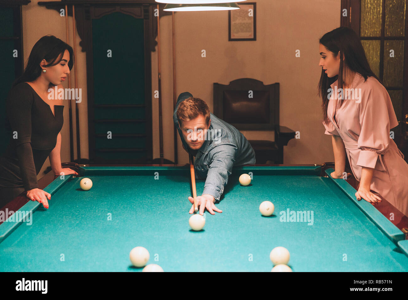Young redhead player stand between two models. He aims into billiard ball. Young women stand and look at gamel. They lean to table Stock Photo