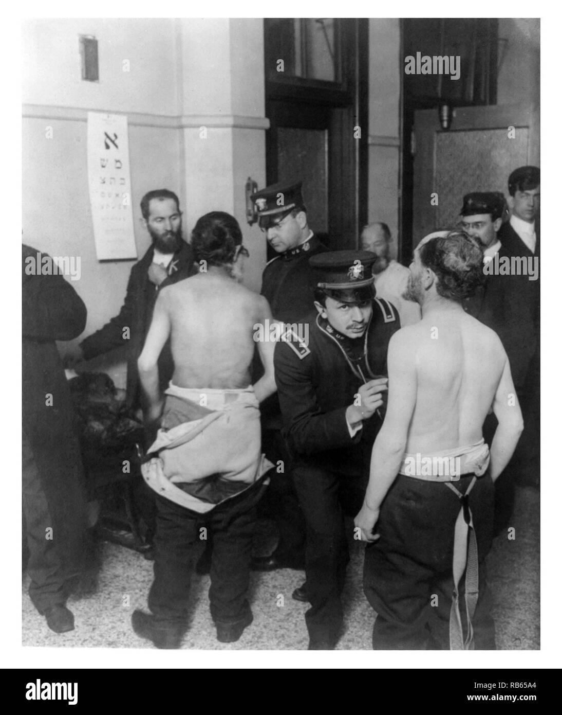 Physicians examining a group of Jewish immigrants who are gathered in small room. Two of the patients have their shirts off and are being examined by physicians while an eye chart written in Hebrew hangs on the wall - possibly taken on Ellis Island. Stock Photo