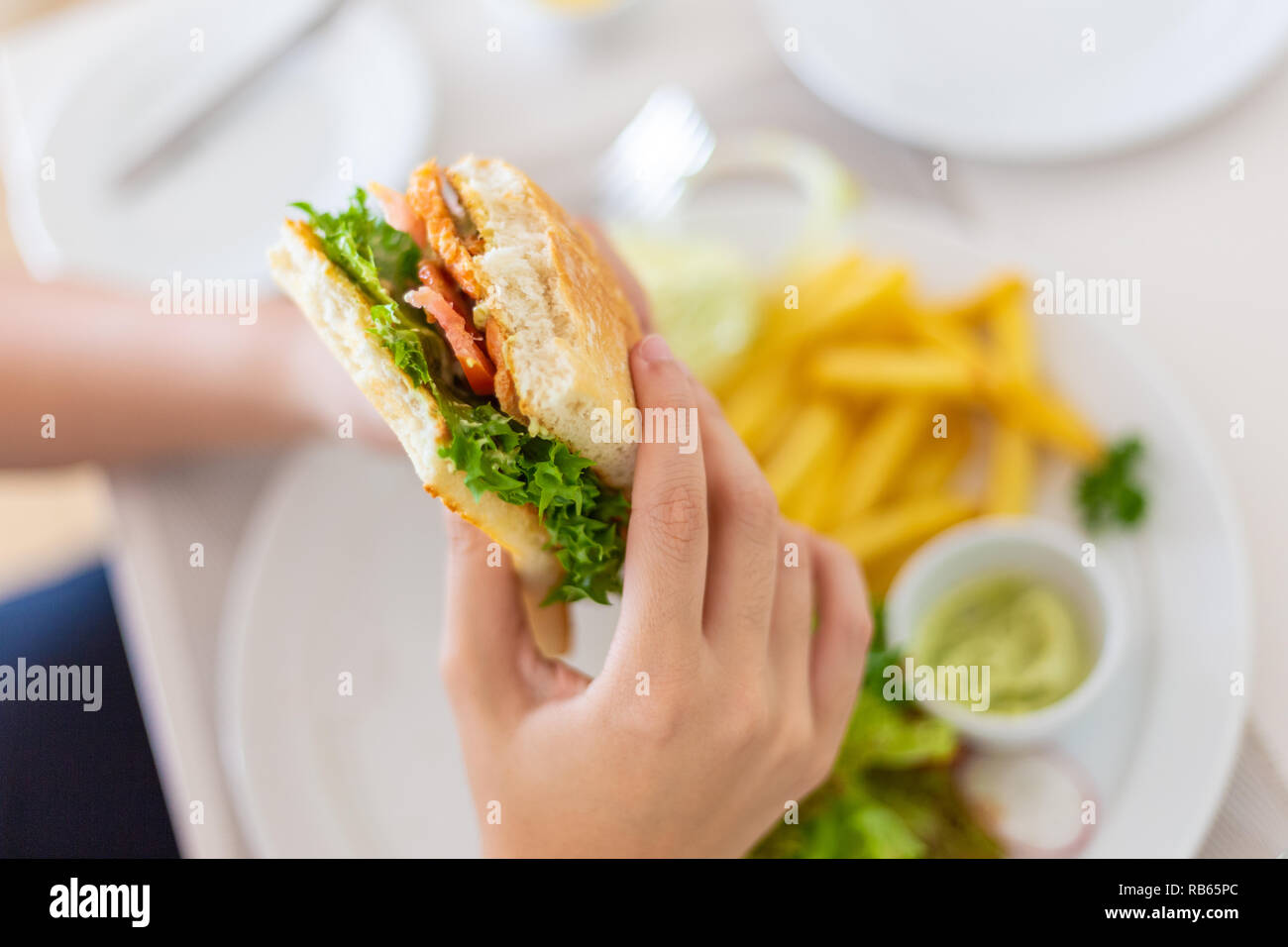 Young female hand holding her half eaten salmon sandwich with the blurred background of french fries,  good for fast food or health concept Stock Photo