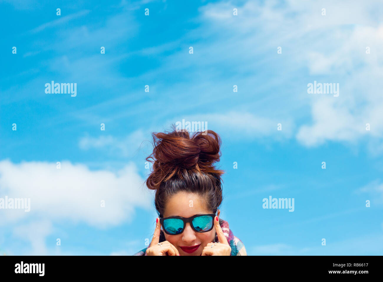 brunette girl with blue glasses and a big messy bun, red lips and nails with the sky as background Stock Photo