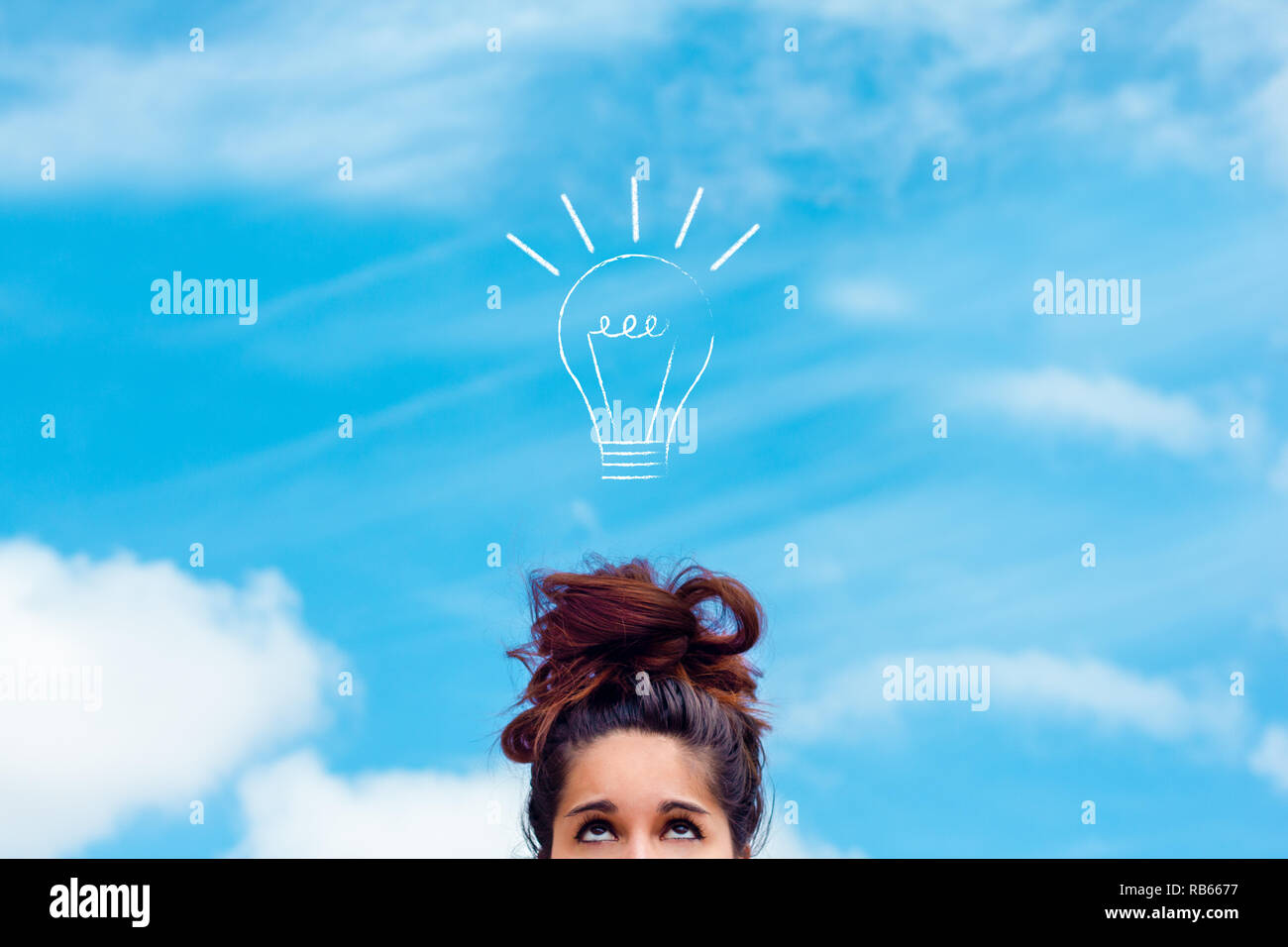 concept of innovation, idea, creativity. Brunette girl with messy bun looking up at a drawn light bulb on the blue sky Stock Photo
