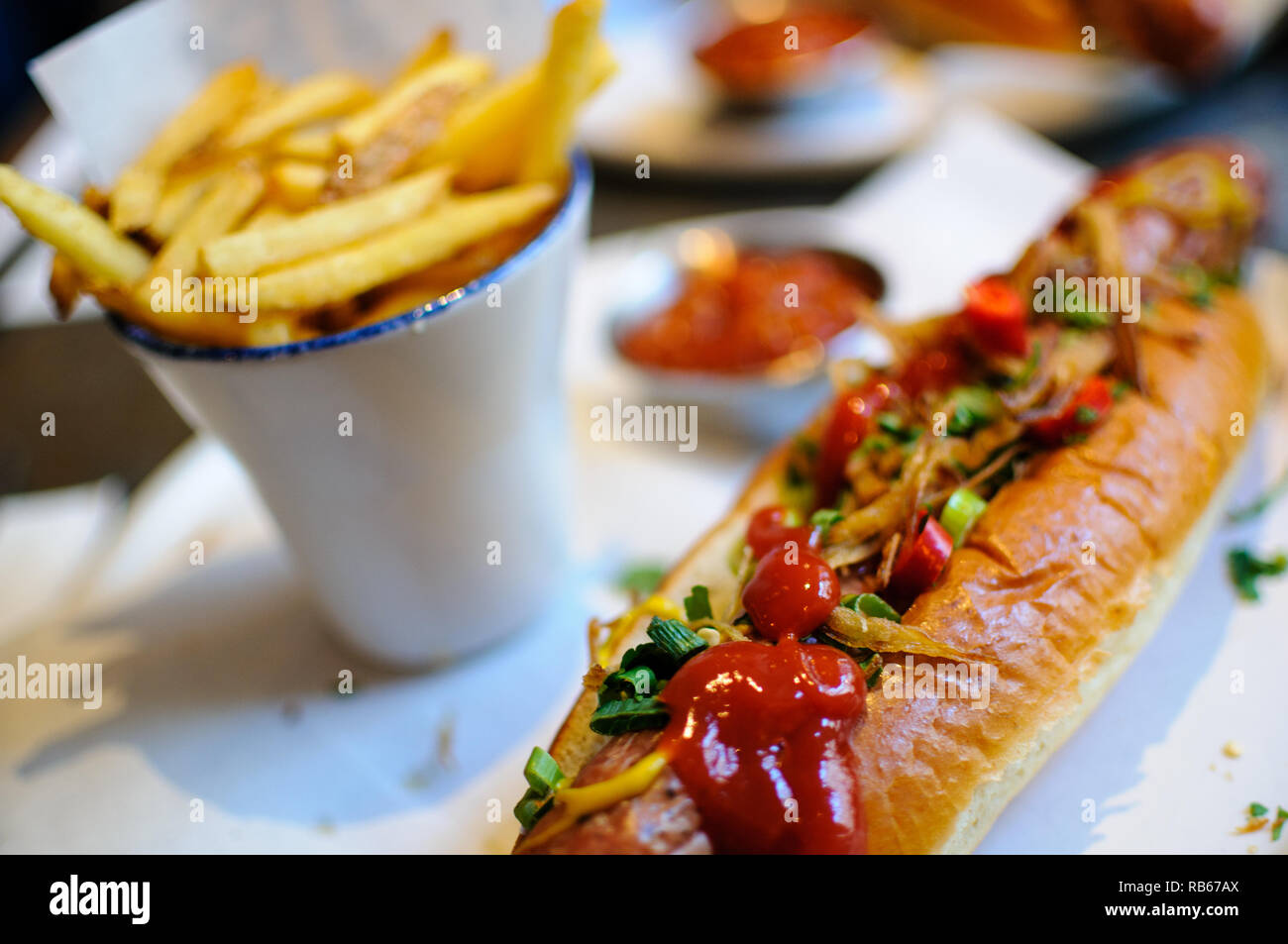 A close-up of a classic hot dog covered in tomato ketchup, mustard, crispy onions and chives with fries / chips in the background softly out of focus. Stock Photo