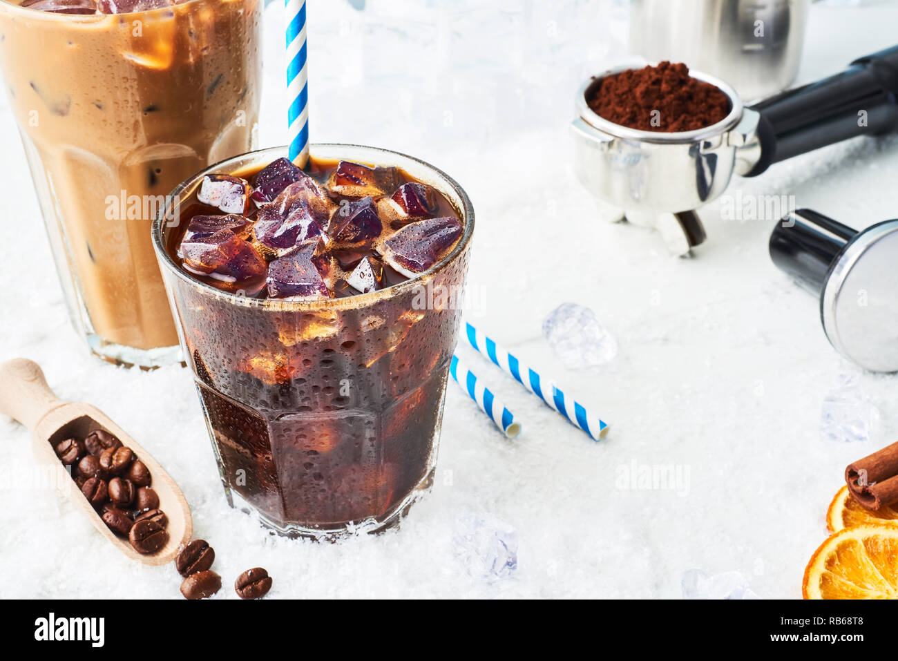 Summer drink iced coffee or soda in a glass and ice coffee with cream in a tall glass surrounded by ice, coffee beans, portafilter, tamper, milk jug a Stock Photo