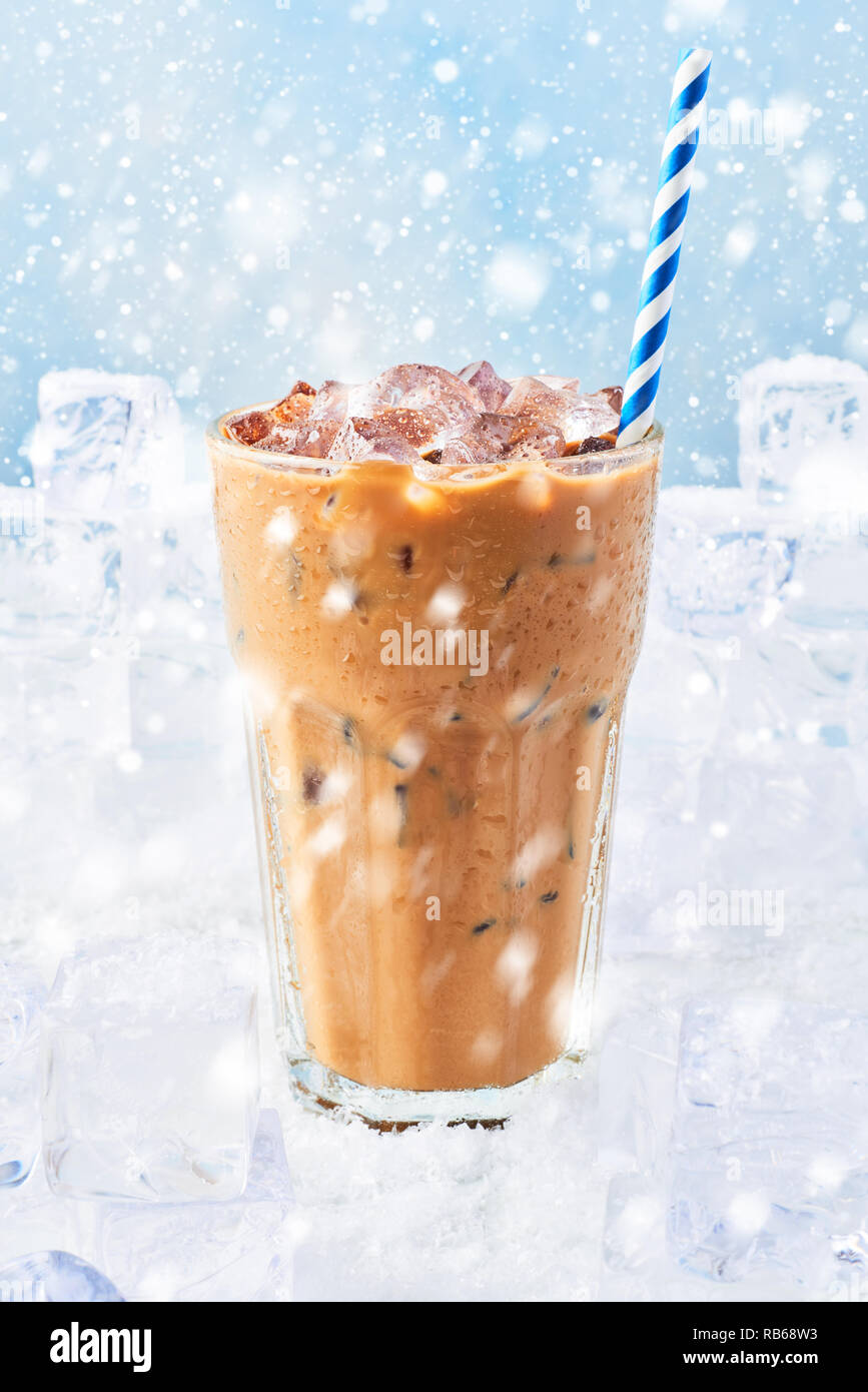 Winter drink ice coffee with cream in a tall glass with straw surrounded by ice cubes on snow over blue background with snow. Selective focus, copy sp Stock Photo