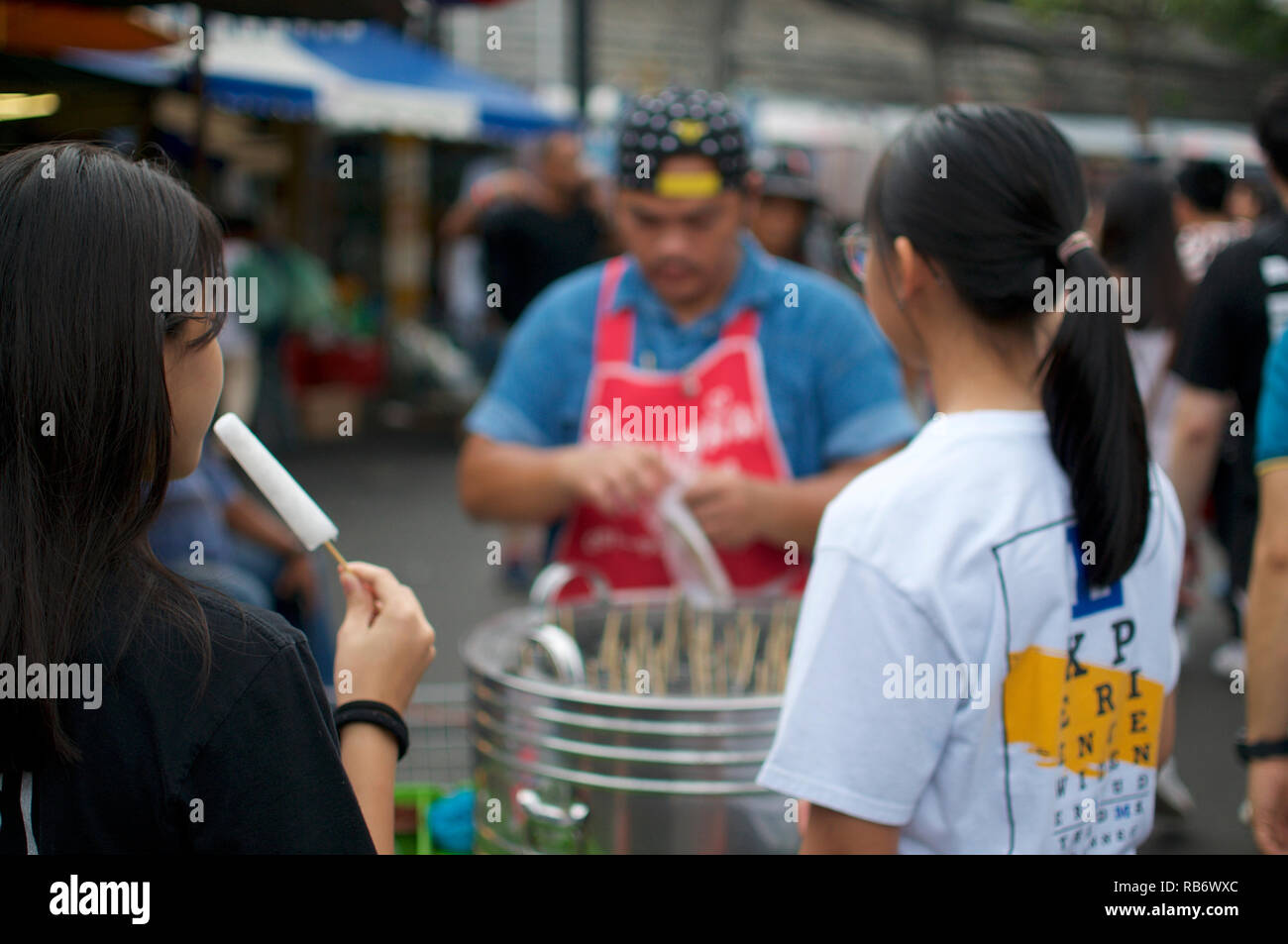 A young woman is eating a Thai popsicle while her friend is waiting for hers in front of the vendor stall at the Chatuchak market, the biggest and most fa Stock Photo