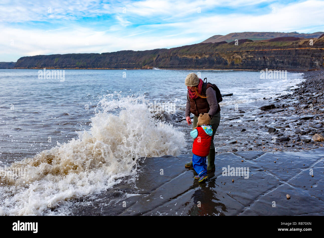 Candid portrait photograph of Parent and young girl standing on coastal ledge getting dredged by crashing waves on Kimmeridge beach, Dorset. Stock Photo