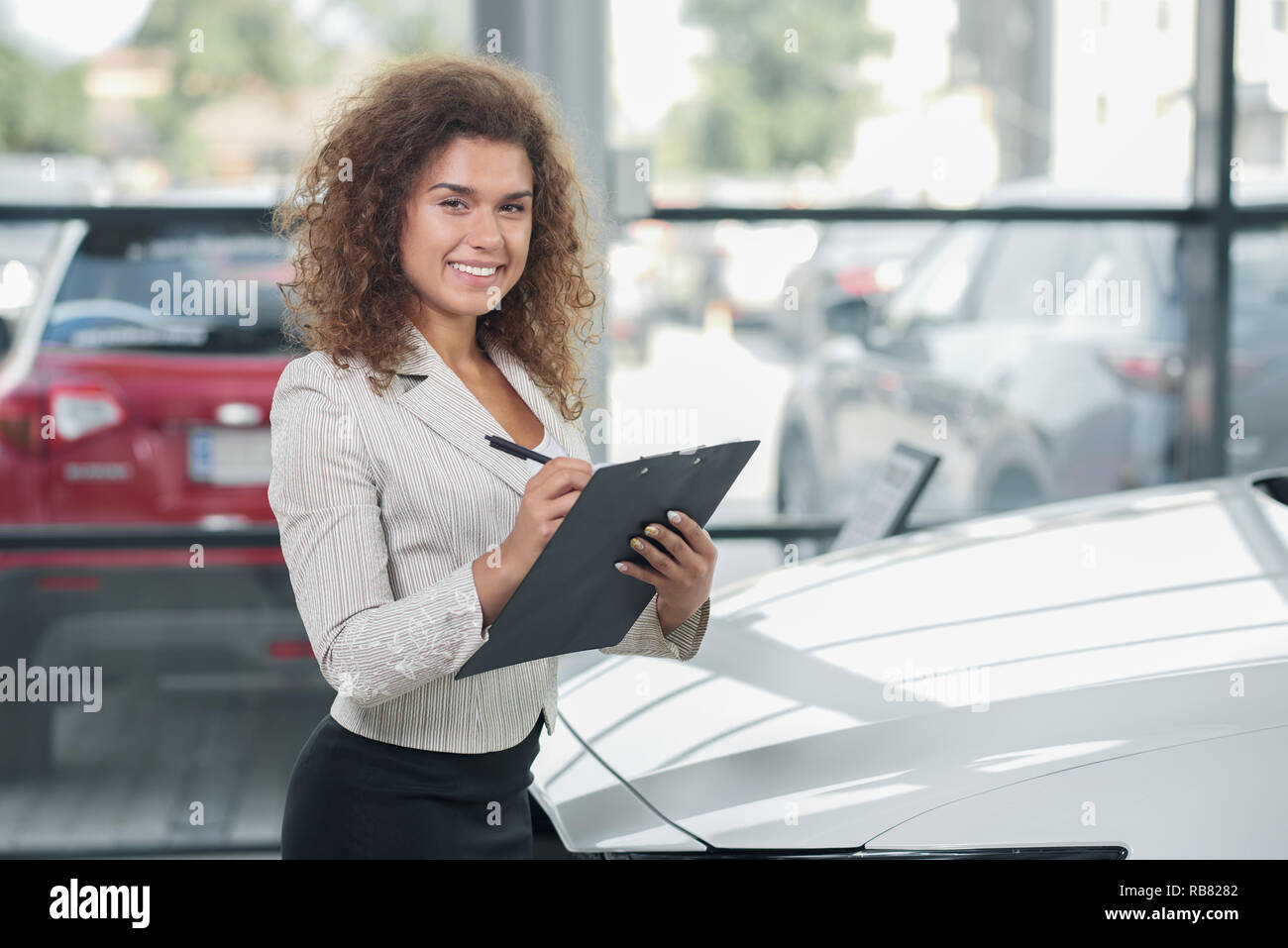Female manager of car dealership standing in showroom and posing. Pretty woman with curly hair smiling, looking at camera. Car dealer holding black folder and pen. Stock Photo