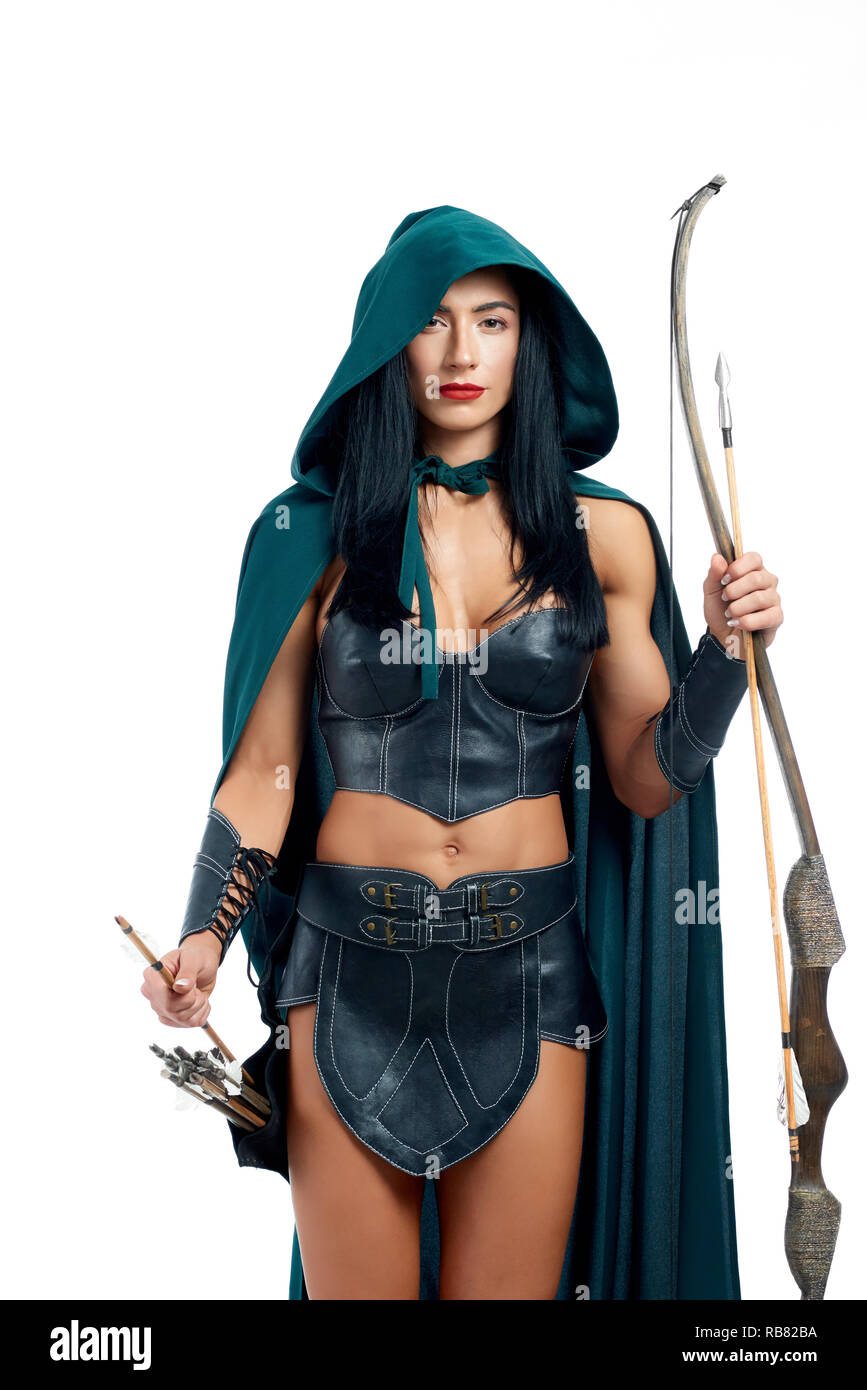 Beautiful and young girl making cosplay of fantasy creature. Gorgeous archery in emerald mantle with hood and leather armor posing at camera. Elegant woman holding bow and arrow and ready for fight. Stock Photo