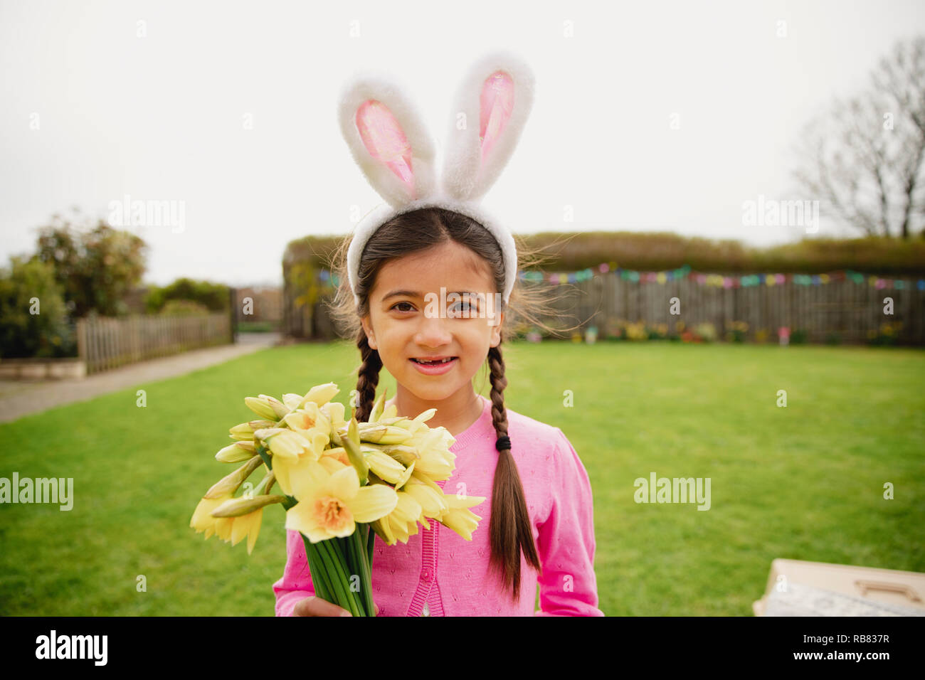 Portrait of a young girl standing outdoors while holding a bunch of daffodils and wearing rabbit ears. Stock Photo