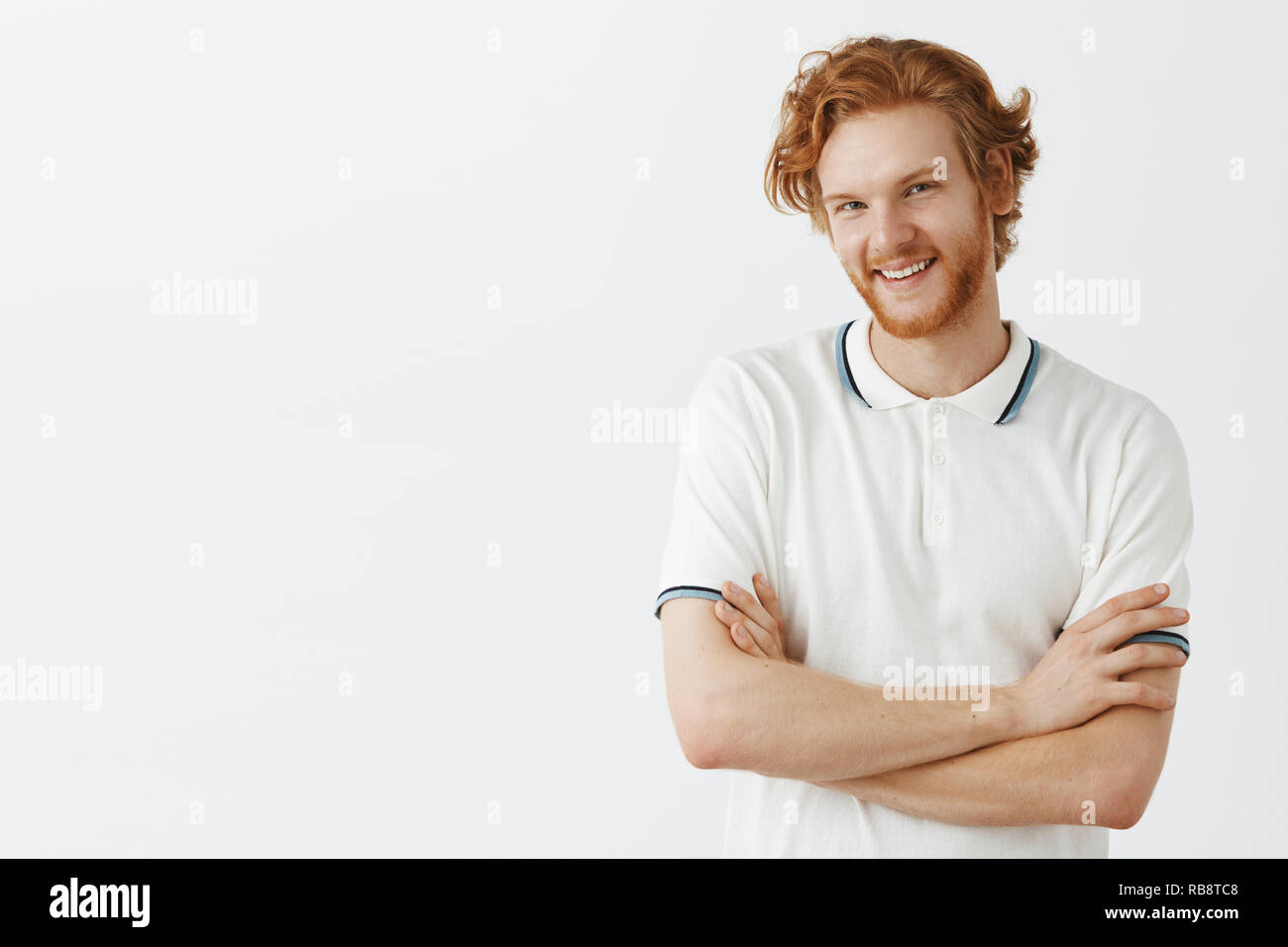 Waist-up shot of cute european redhead guy with wavy hair and beard tilting head joyfull holding hands crossed on chest and raising one shoulder as if flirting or having romantic mood Stock Photo