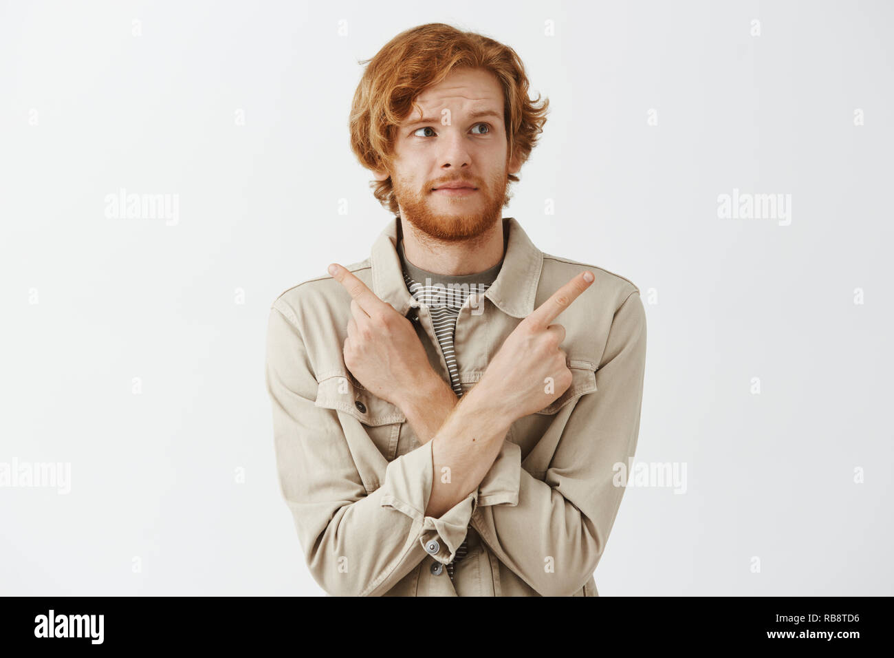 Studio sht of confused cute redhead european male model with sick beard in jacket over t-shirt pointing with crossed hands in different directions both left and right being questioned where to go Stock Photo