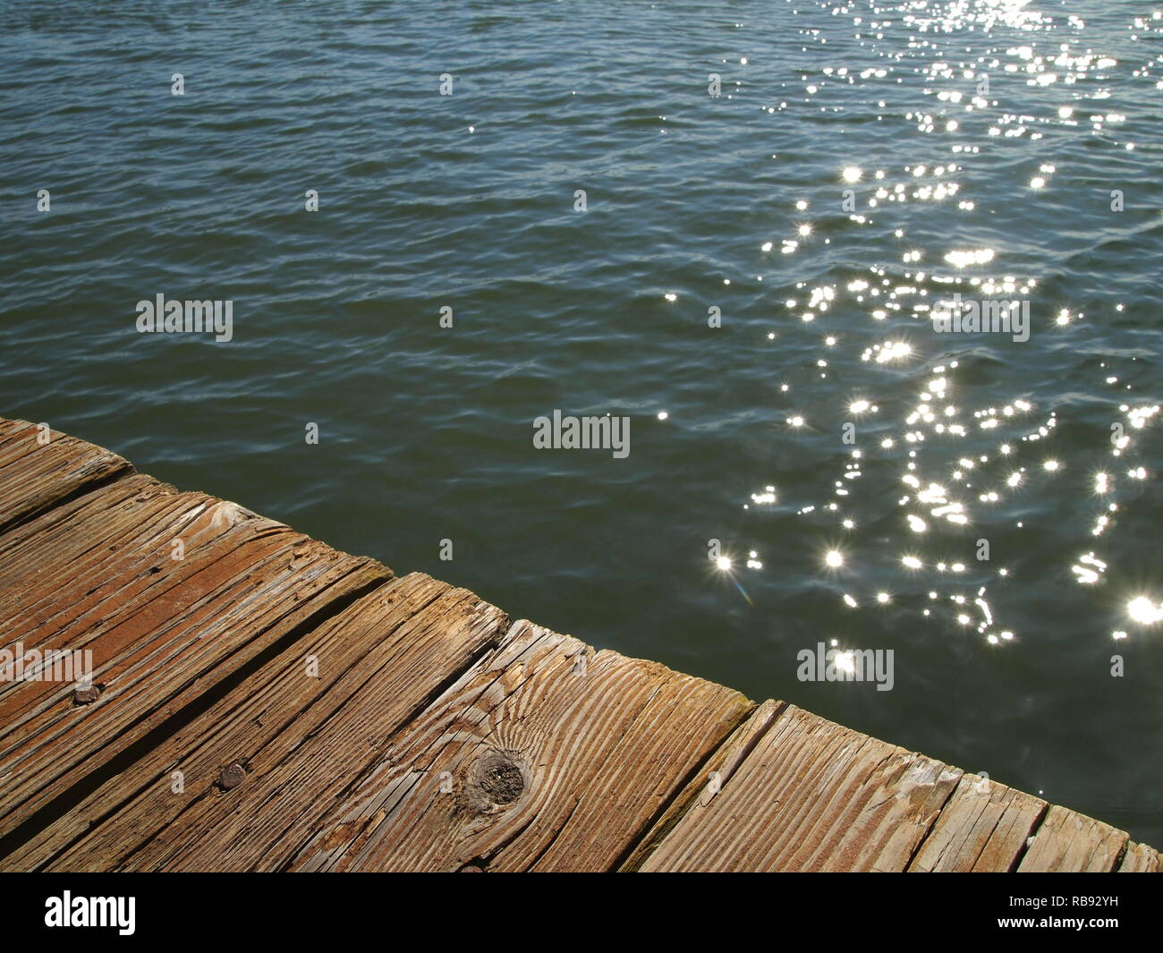Glistening sunlight reflections on water beneath wooden dock. Close-up perspective of weathered wooden boards. Angled perspective, minimalism, texture Stock Photo