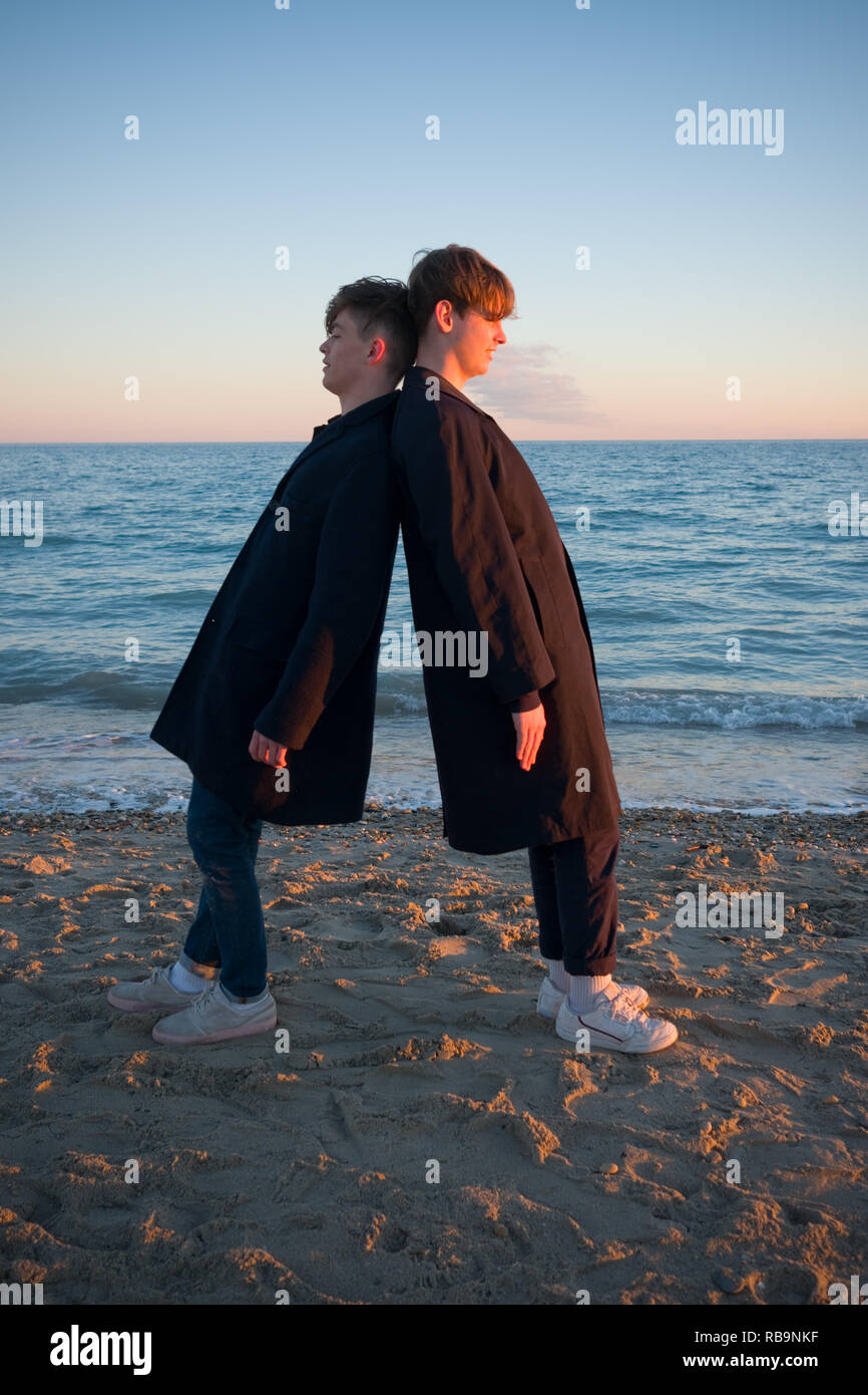 two teenage boys wearing similar clothes fool about on a beach by leaning against each other as the sunsets in southern france Stock Photo