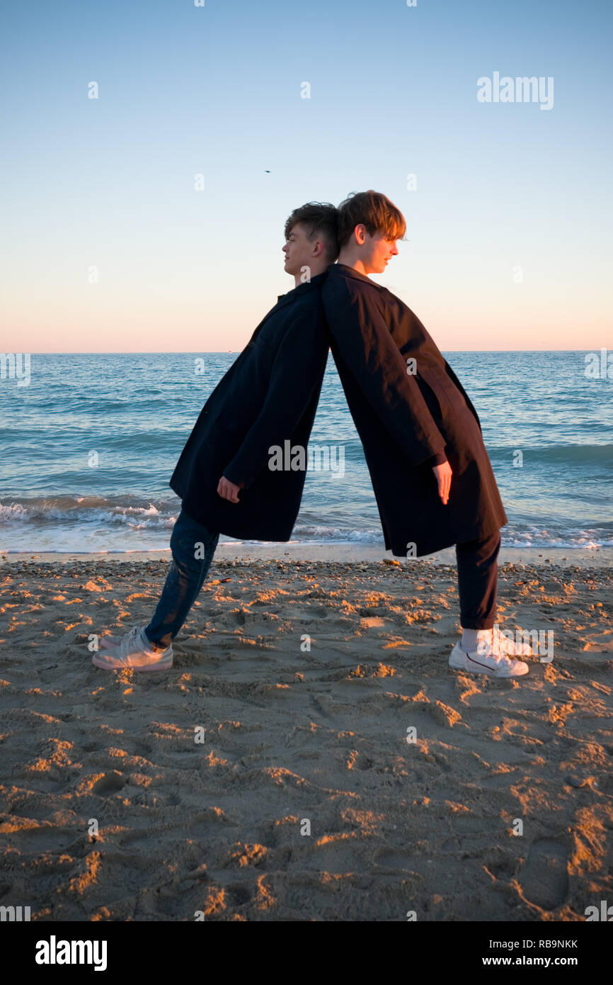 two teenage boys wearing similar clothes fool about on a beach by leaning against each other as the sunsets in southern france Stock Photo
