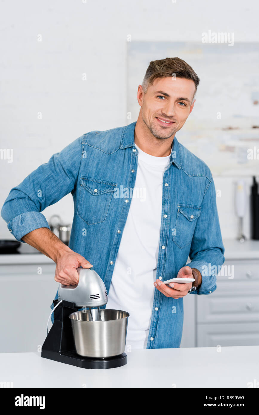 Man Using Kitchen Machine adult man using kitchen mixer and smartphone while smiling at camera ...