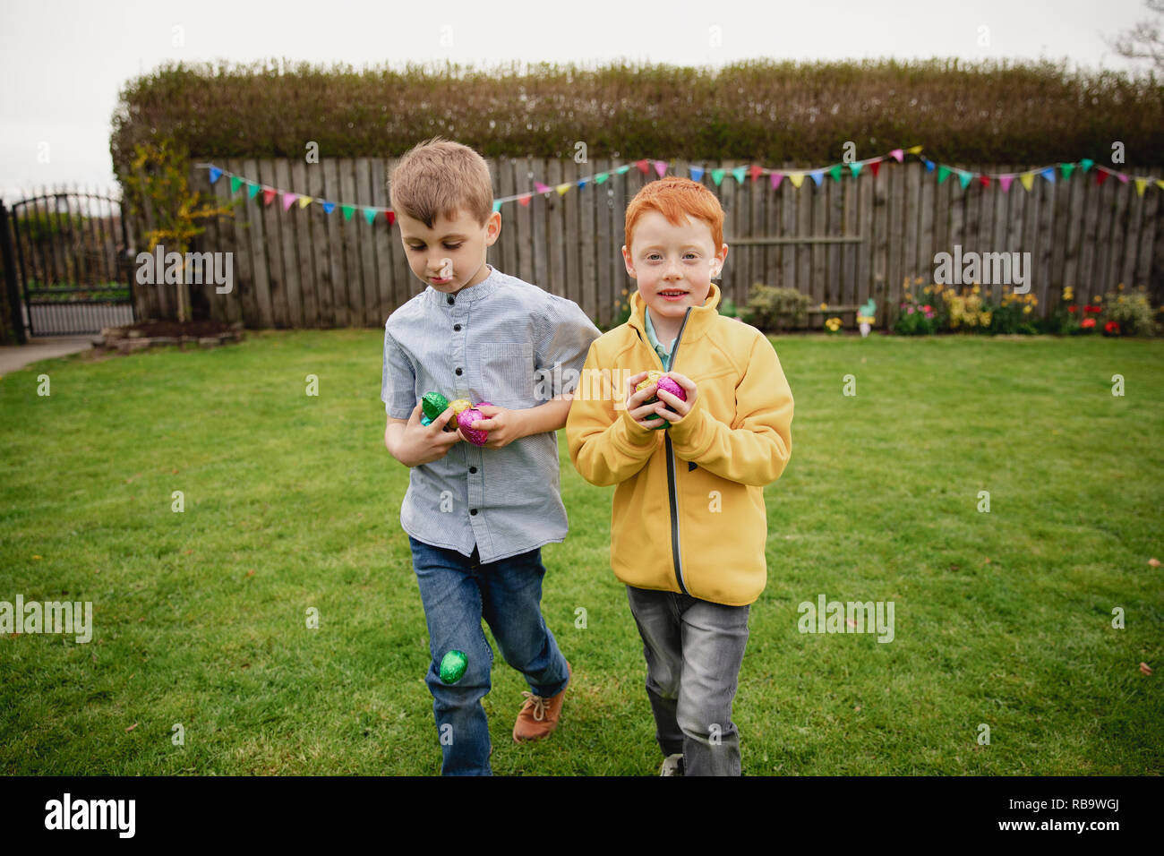 Two young boys walking through the back garden after an easter egg hunt. They are holding handfuls of chocolate eggs in their hands. Stock Photo