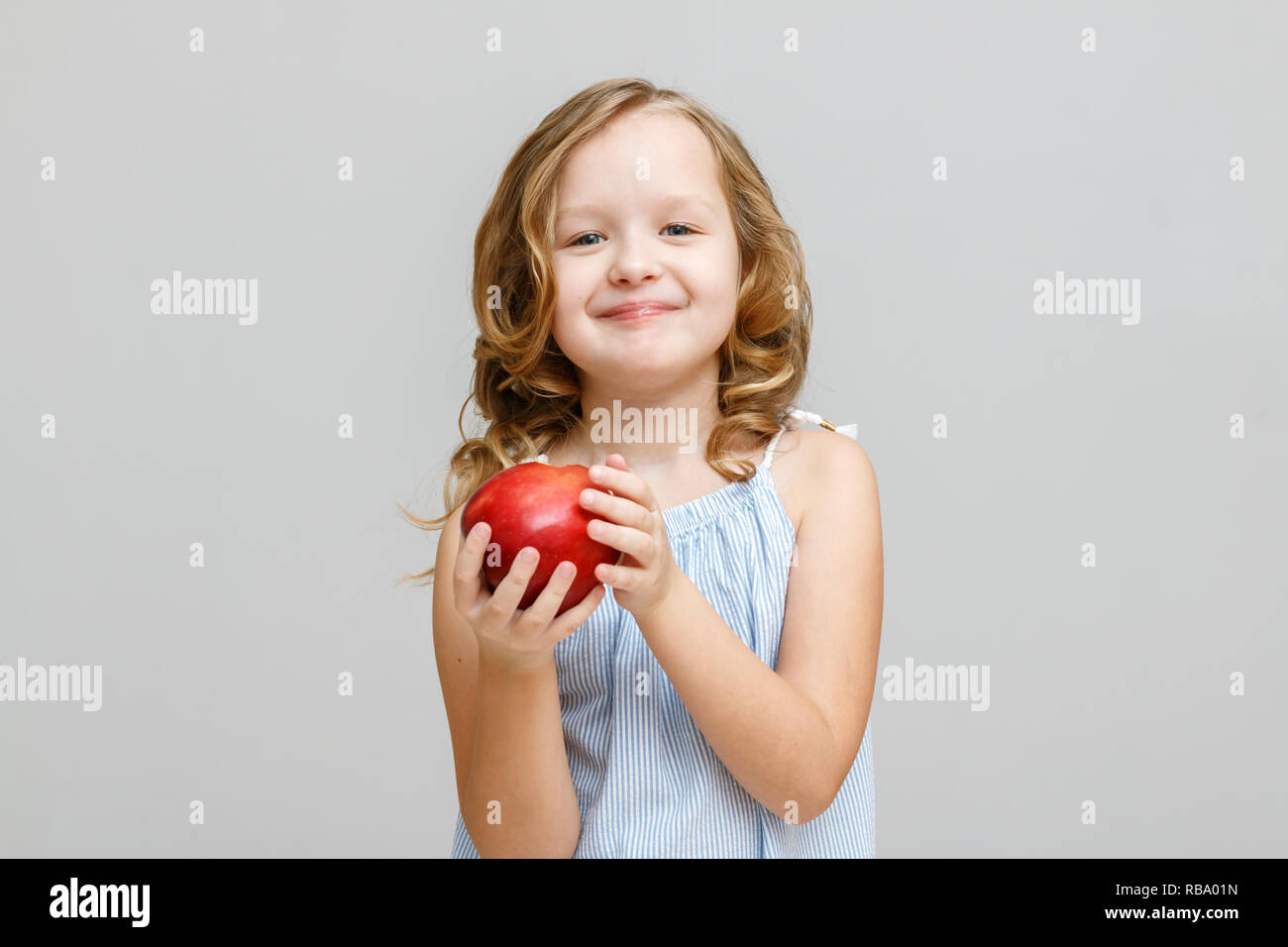 Portrait of a happy smiling little blonde girl on a gray background. Baby eating red apple Stock Photo