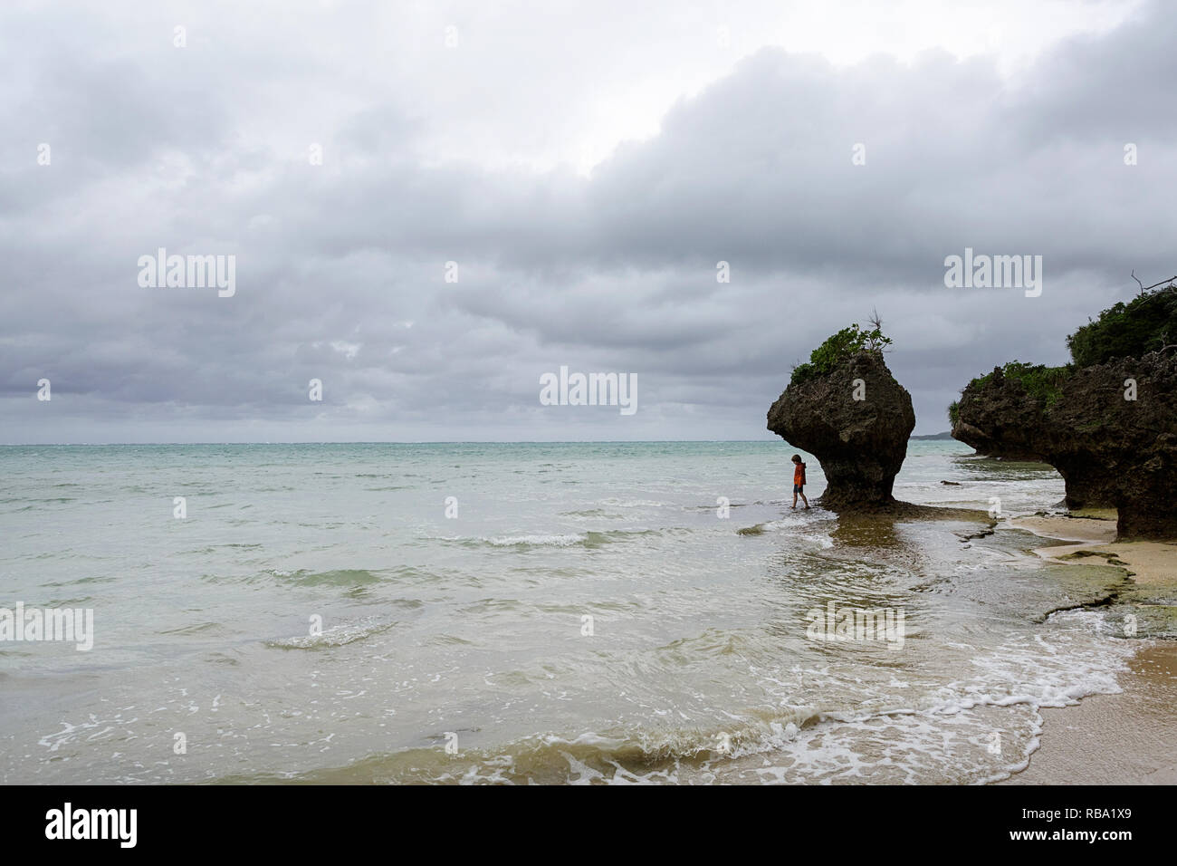 Young boy standing under limestone sculpture by the sea at sandy beach at Ibaruma Sabichi Cave, Ishigaki, Japan Stock Photo
