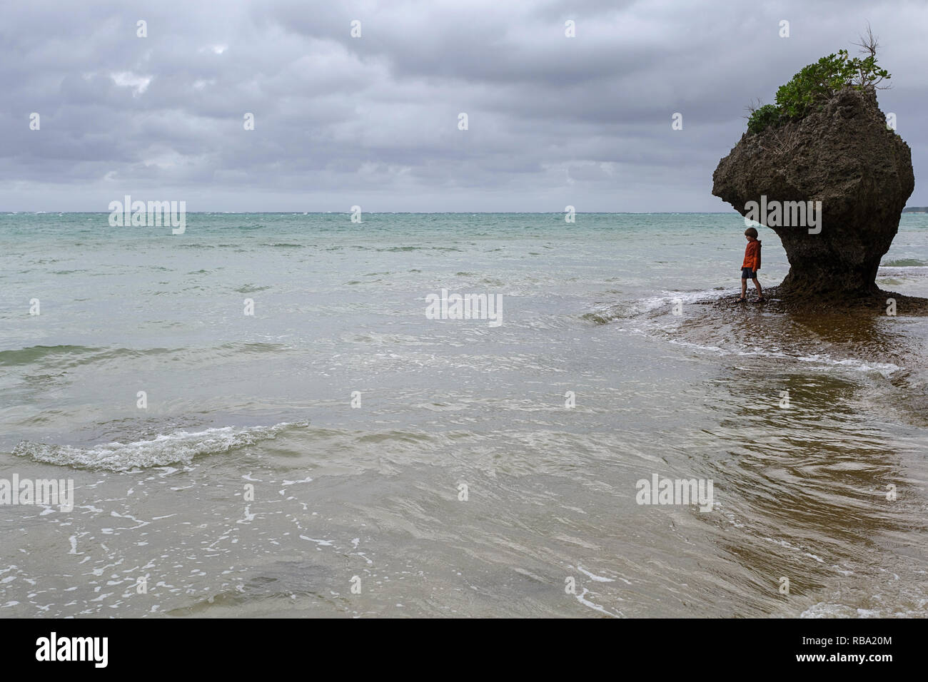 Young boy standing under limestone sculpture by the sea at sandy beach at Ibaruma Sabichi Cave, Ishigaki, Japan Stock Photo